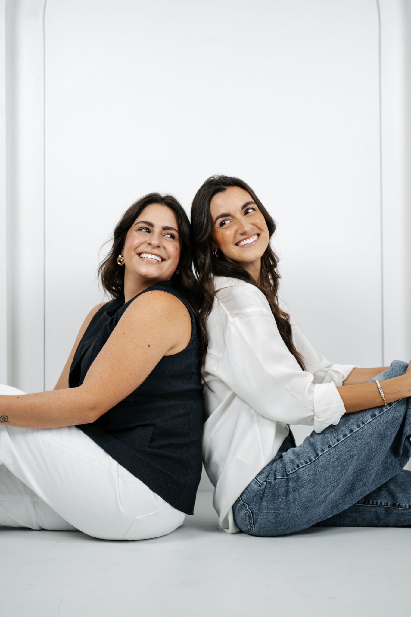 Two friends in casual outfits lying down and laughing together against a white backdrop in a photo studio series.