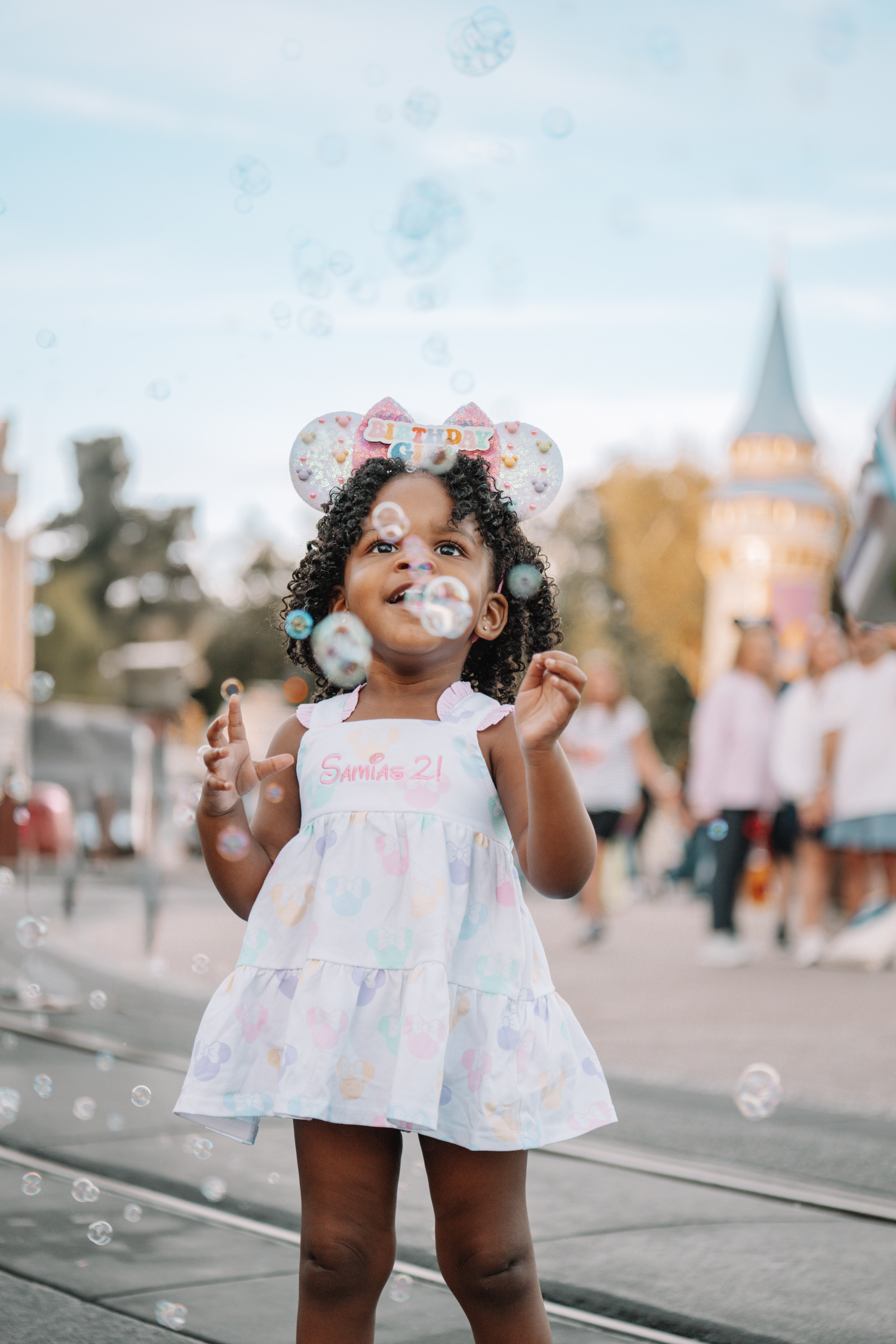 Young child in floral dress plays with bubbles outdoors on sunny day.