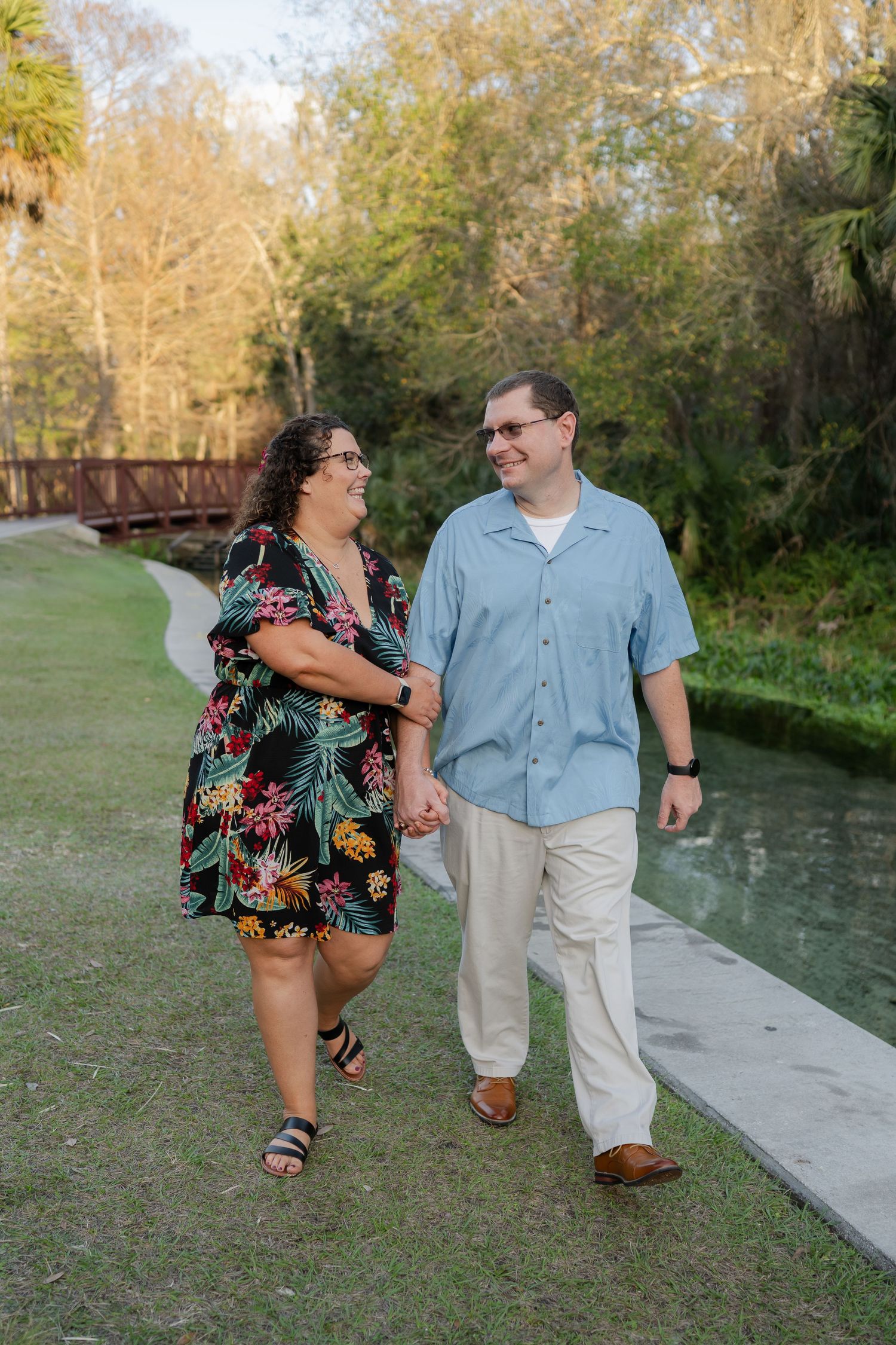 A couple walks together and laughs along a waterfront path surrounded by palm trees.