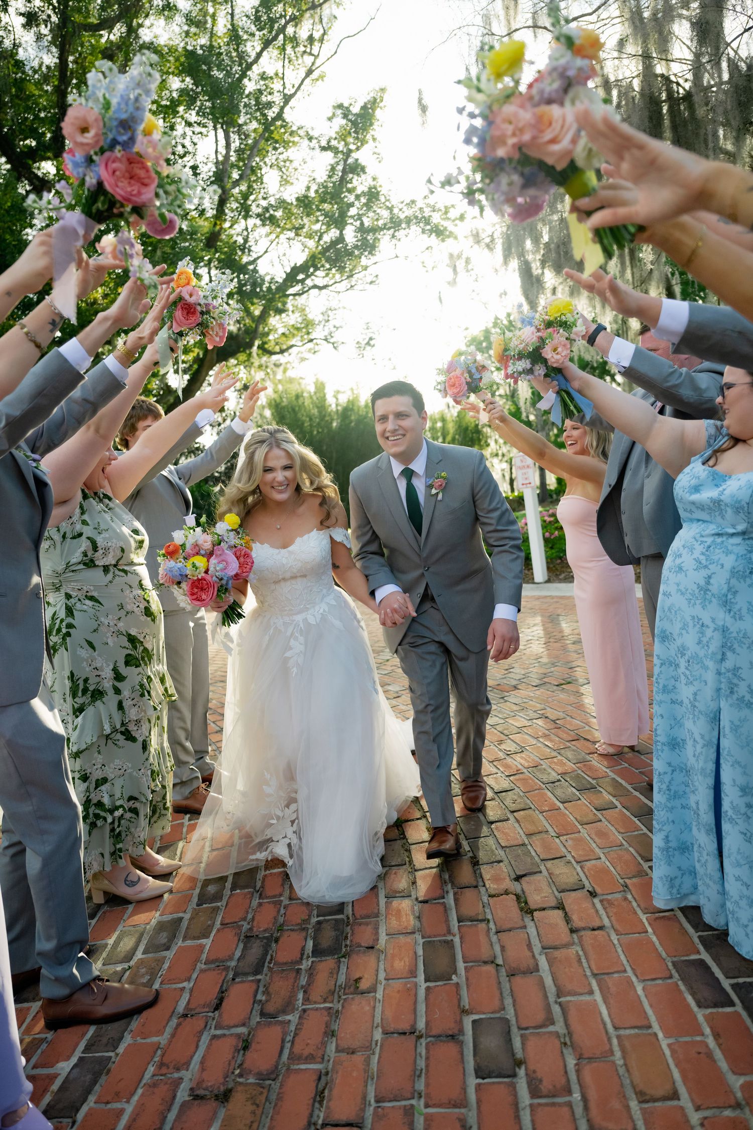 Wedding party celebrates on brick walkway with bridesmaids in blue and pink dresses throwing flower petals in joyful sequence.