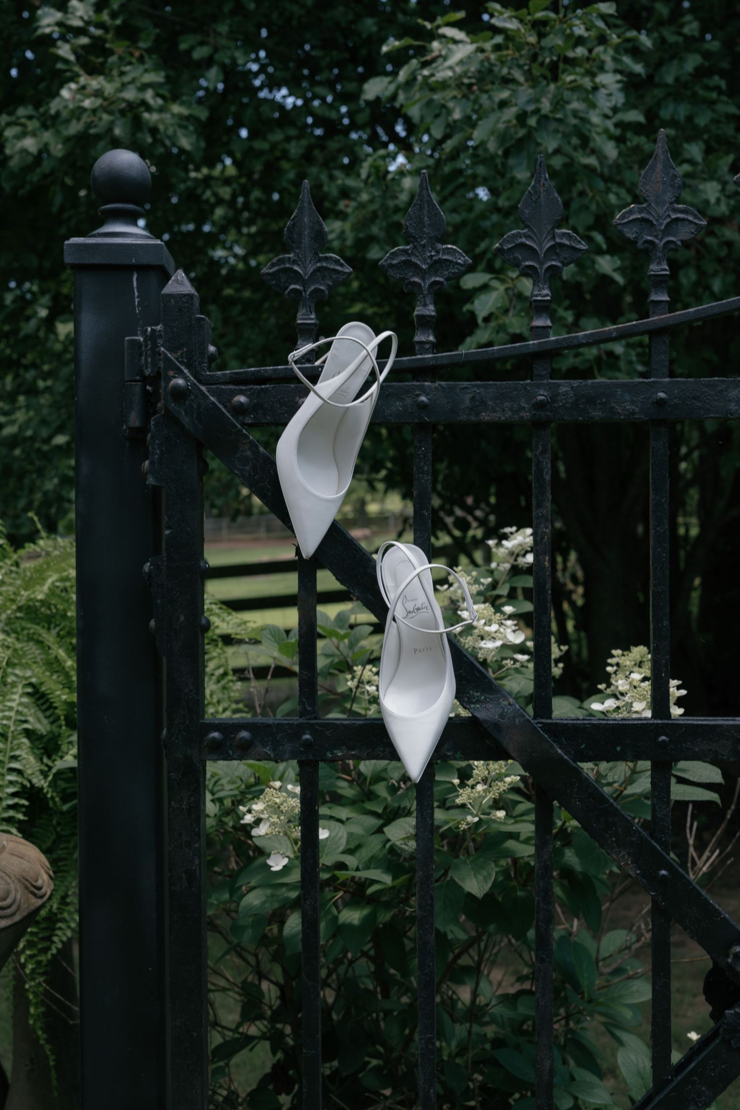 White bridal heels displayed on a black ornate iron garden gate surrounded by greenery.