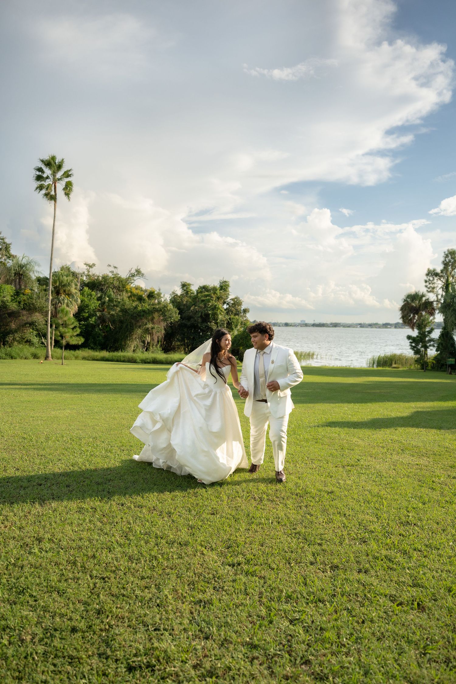 Bride in a flowing white gown and groom in a white suit walk hand in hand across a sunny lakeside lawn under a bright cloudy sky.