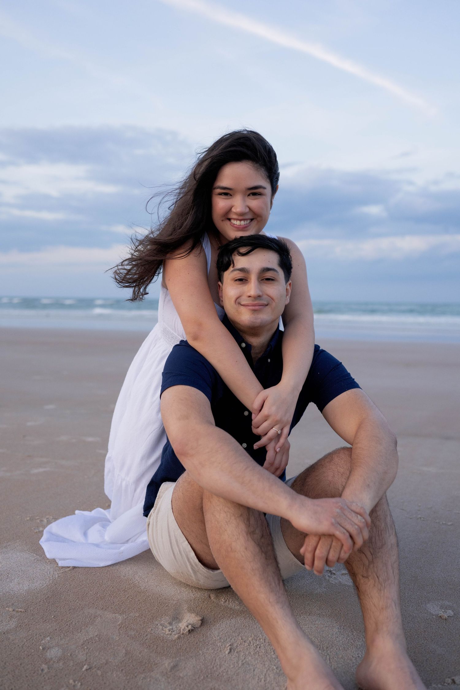 A smiling couple poses on a sandy beach at dusk, woman in white dress hugging man seated on sand.