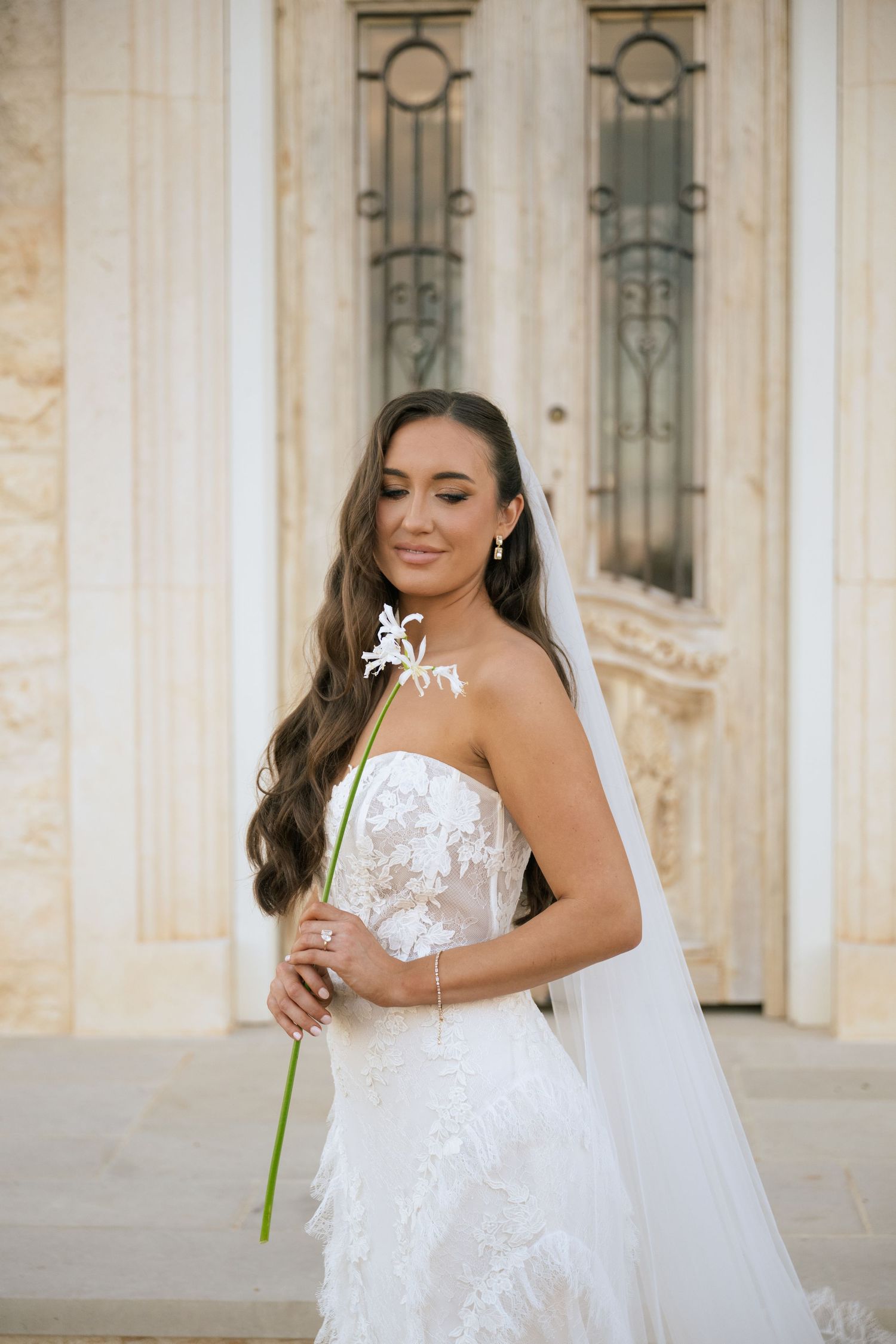 Bride in strapless lace gown with feather hem holds white flower, wearing veil, posing before marble building.