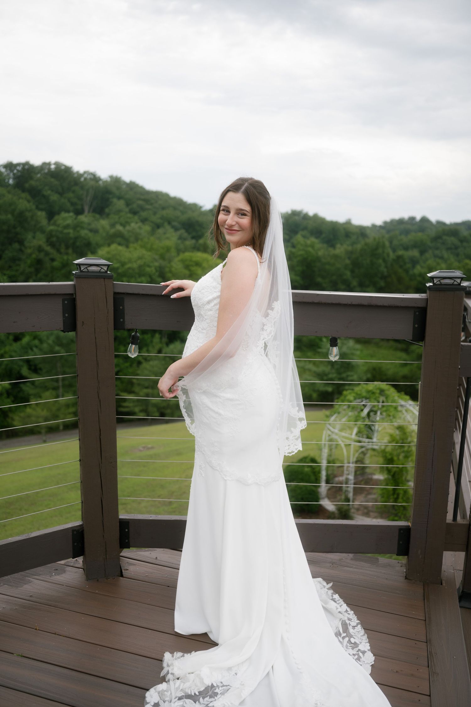 Bride in white wedding gown with veil poses on outdoor deck overlooking lush green trees on cloudy day.