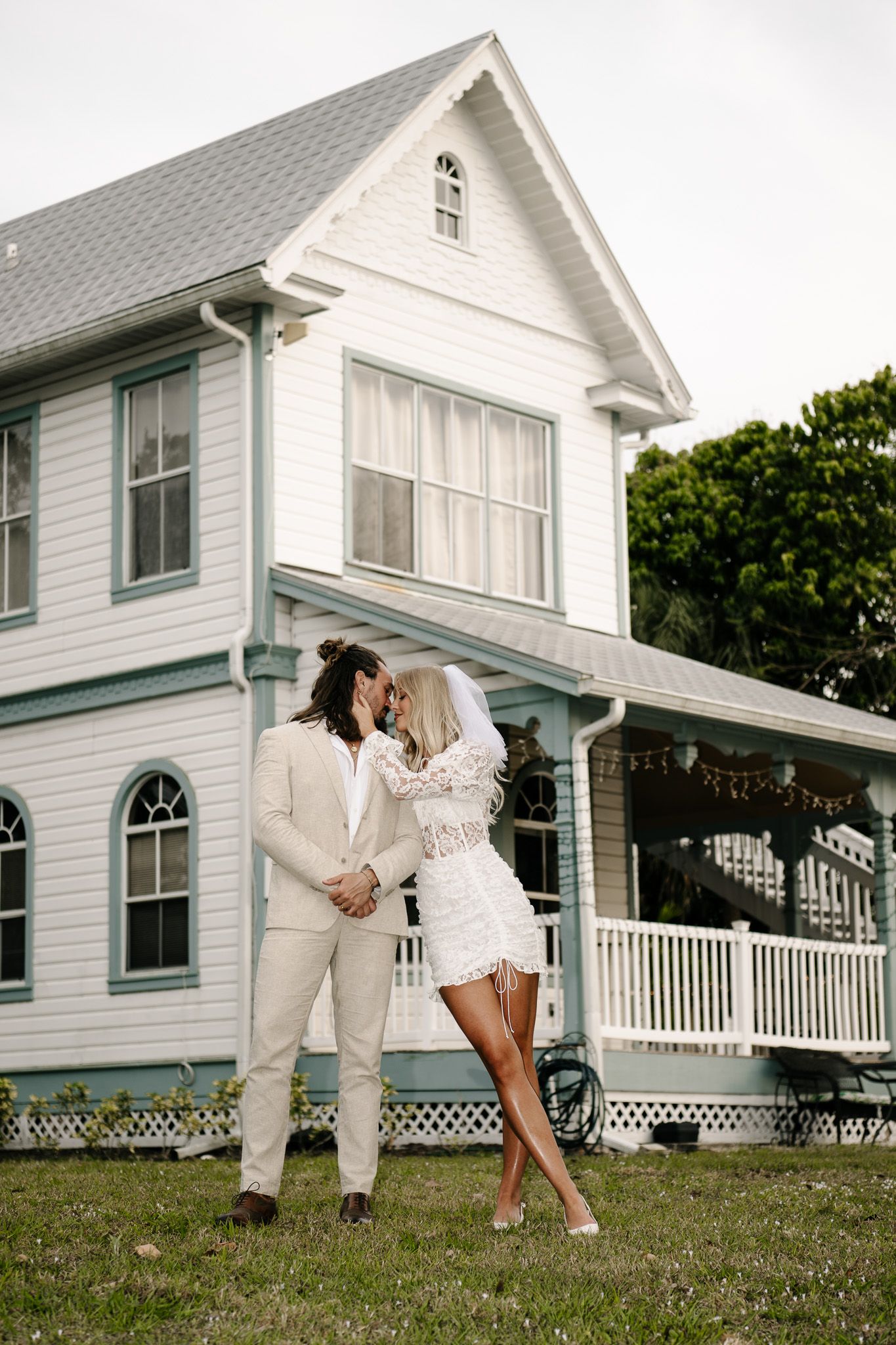 Wedding couple poses dramatically outside white farmhouse with wraparound porch.