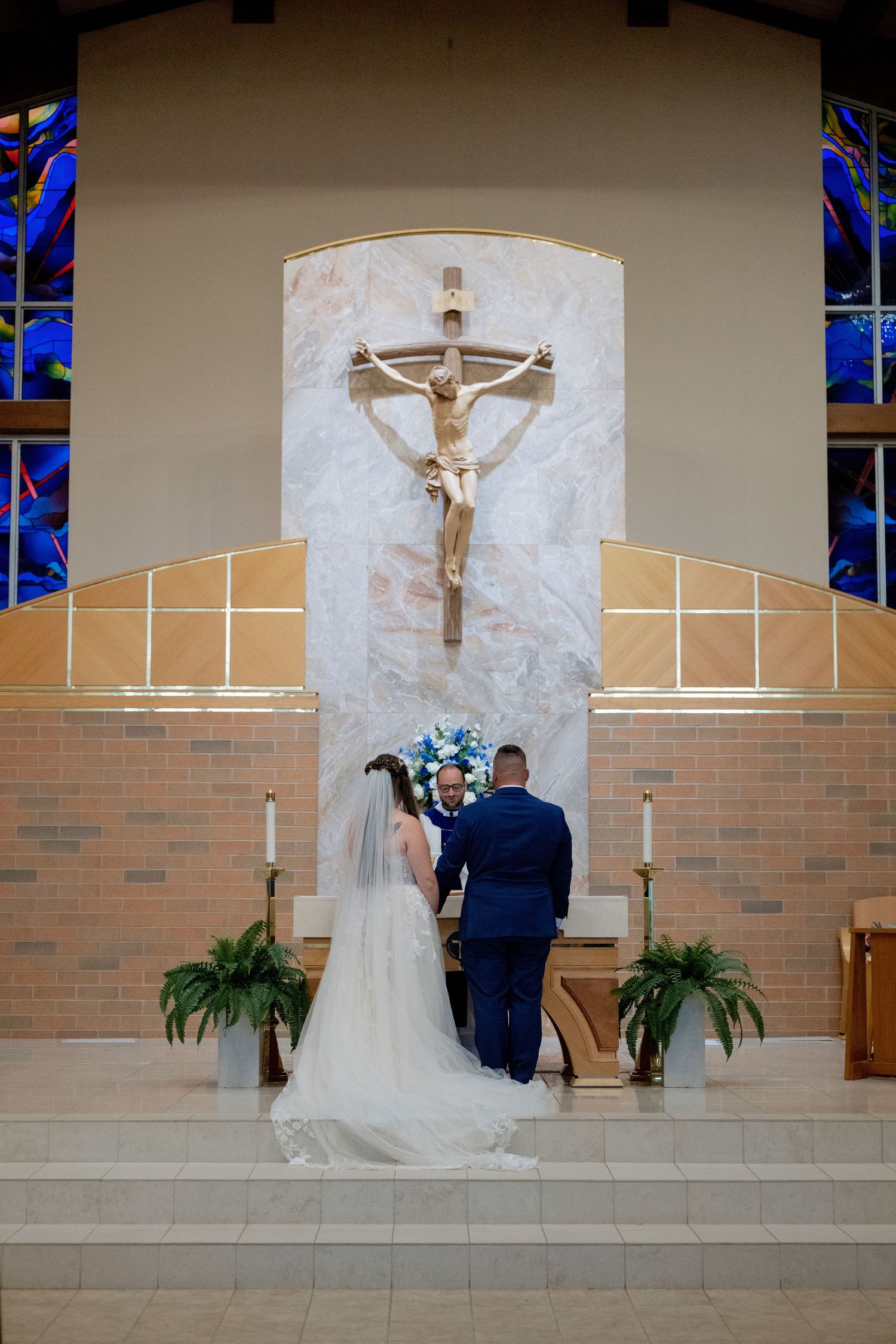 Bride and groom stand at church altar before crucifix during Catholic wedding ceremony with blue floral arrangement.