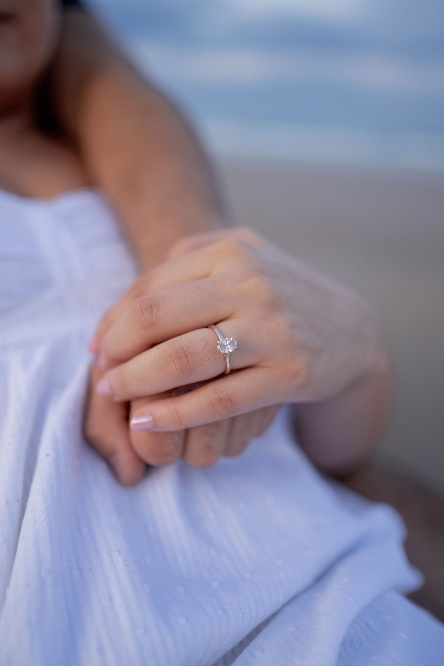 Close-up of a diamond solitaire engagement ring on a woman's hand, wearing a white dress at the beach.