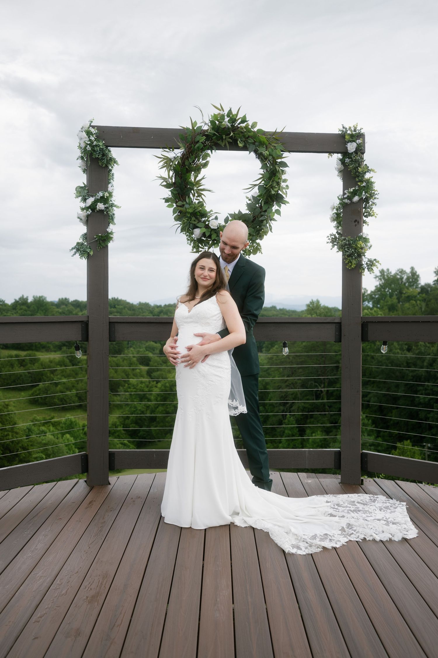 Pregnant bride and groom pose under heart-shaped greenery arch on wooden deck with scenic green landscape backdrop.