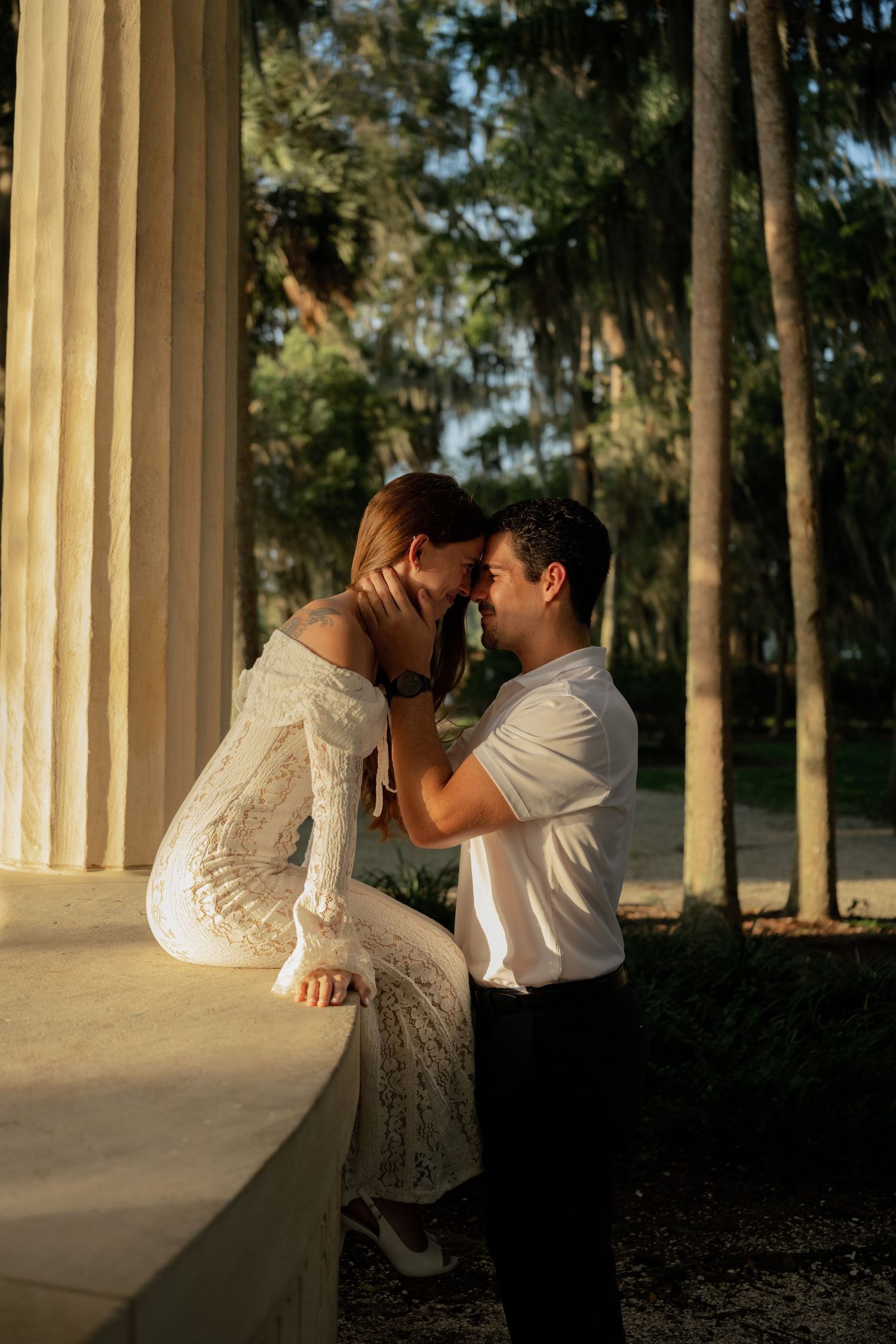 A couple shares a romantic moment near white columns and tall trees, the woman in a white lace dress as the man gently holds her face.
