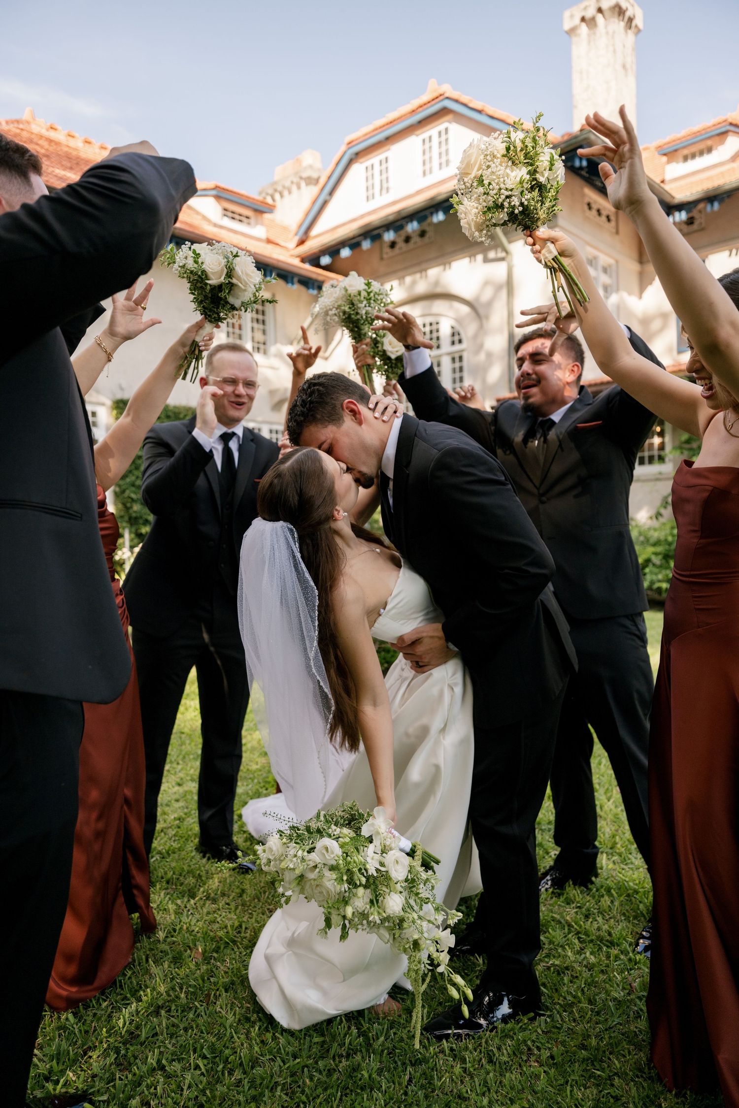 Bride and groom share a dipped kiss on a sunny lawn as joyful wedding guests raise white floral bouquets in front of a historic building.