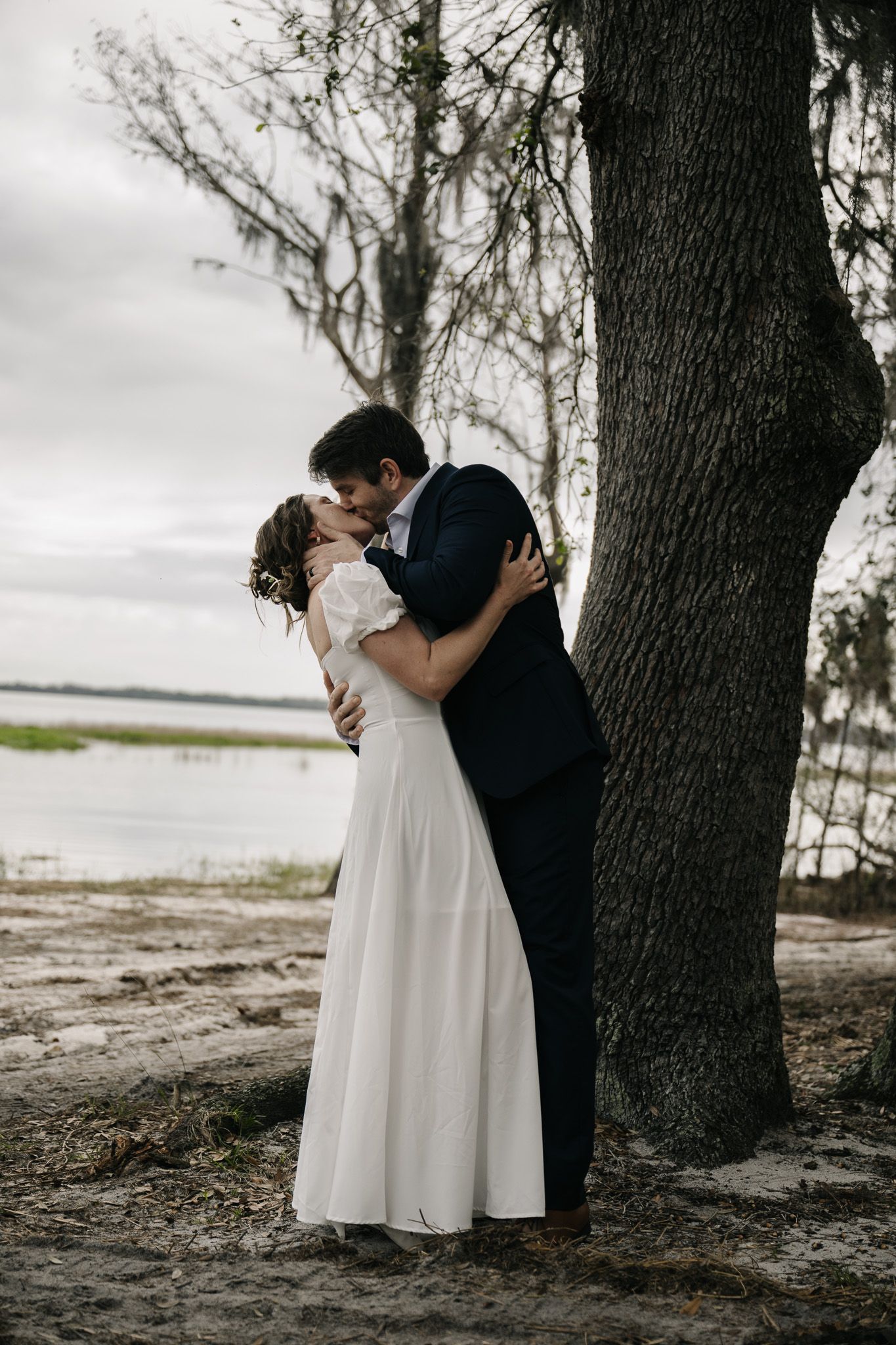 A romantic couple embraces under a large tree by the water at dusk.