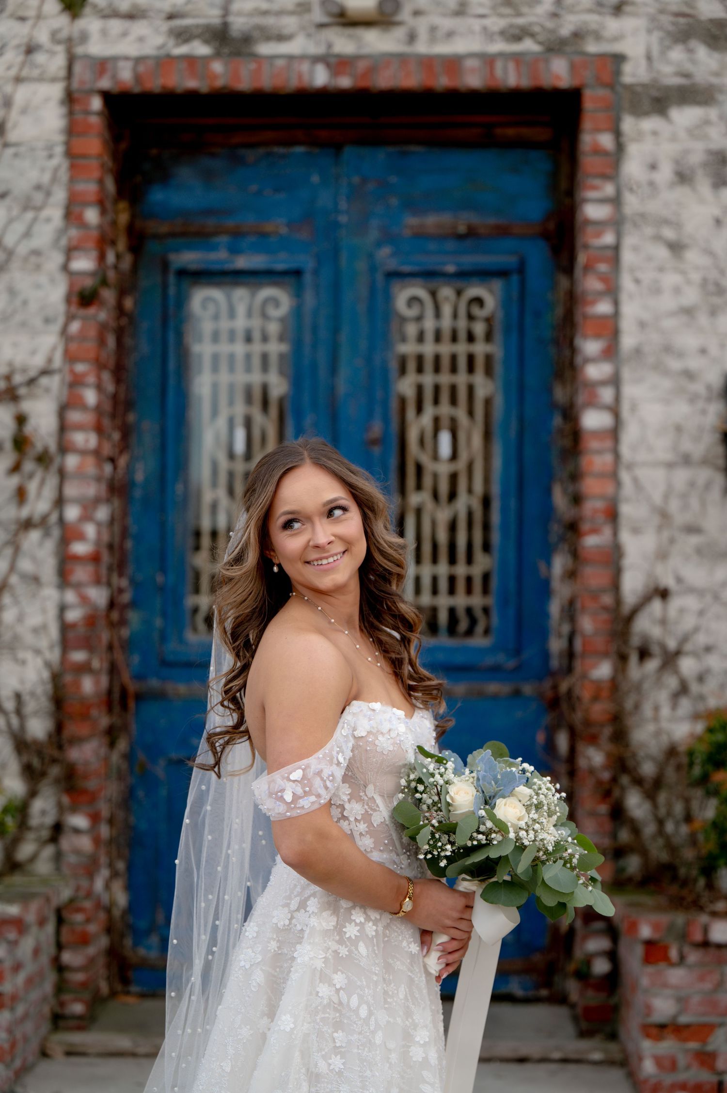 Bride in off-shoulder lace gown holds white bouquet, smiling before rustic blue doors with brick wall backdrop.