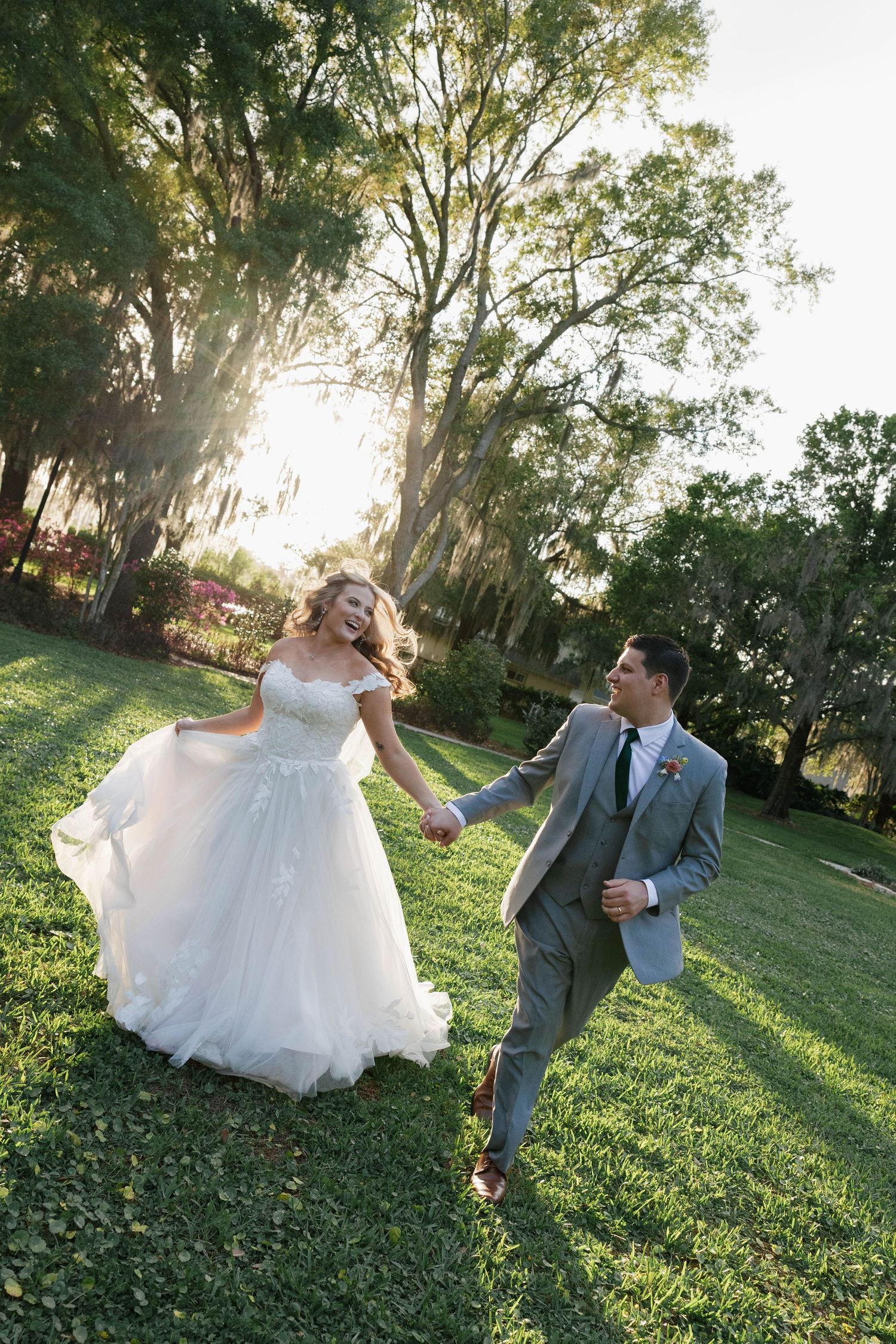 A joyful bride in a white ball gown and groom in a gray suit hold hands running through a sunlit green park.