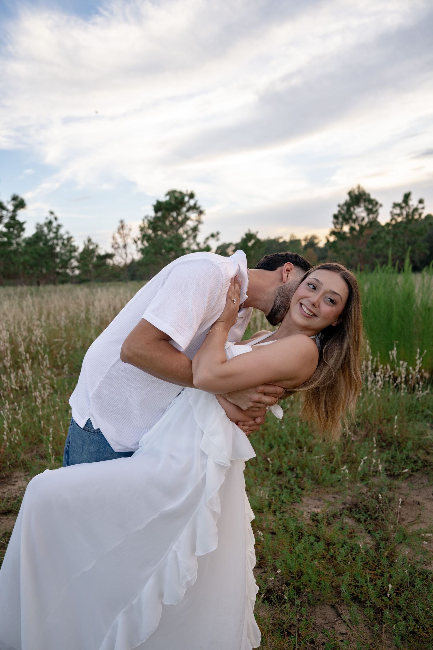 Couple embracing outdoors, man kissing woman's cheek as she smiles, wearing white dress in a green field at dusk.