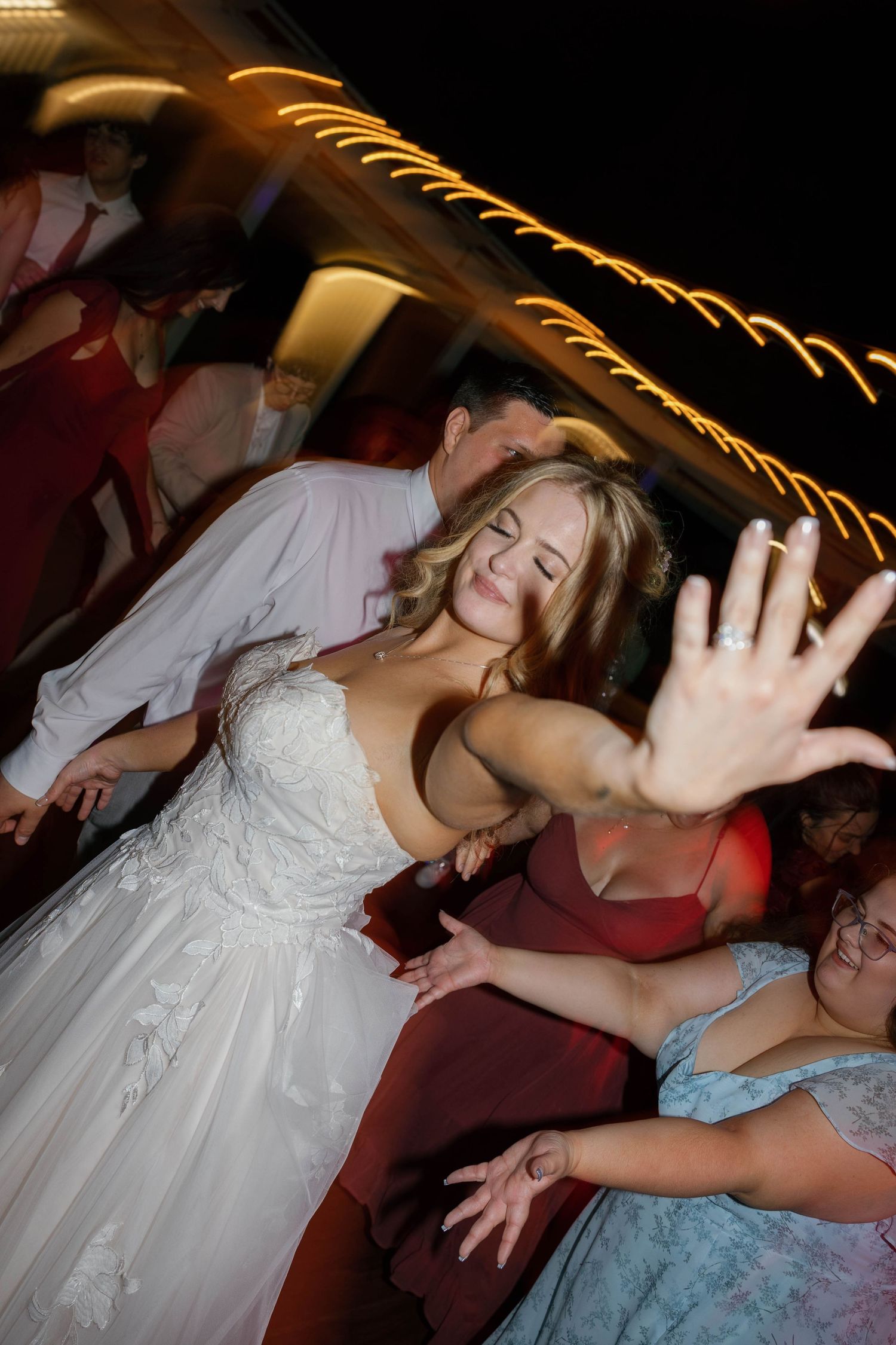 Wedding guests dancing and celebrating on a dimly lit dance floor with decorative string lights overhead.