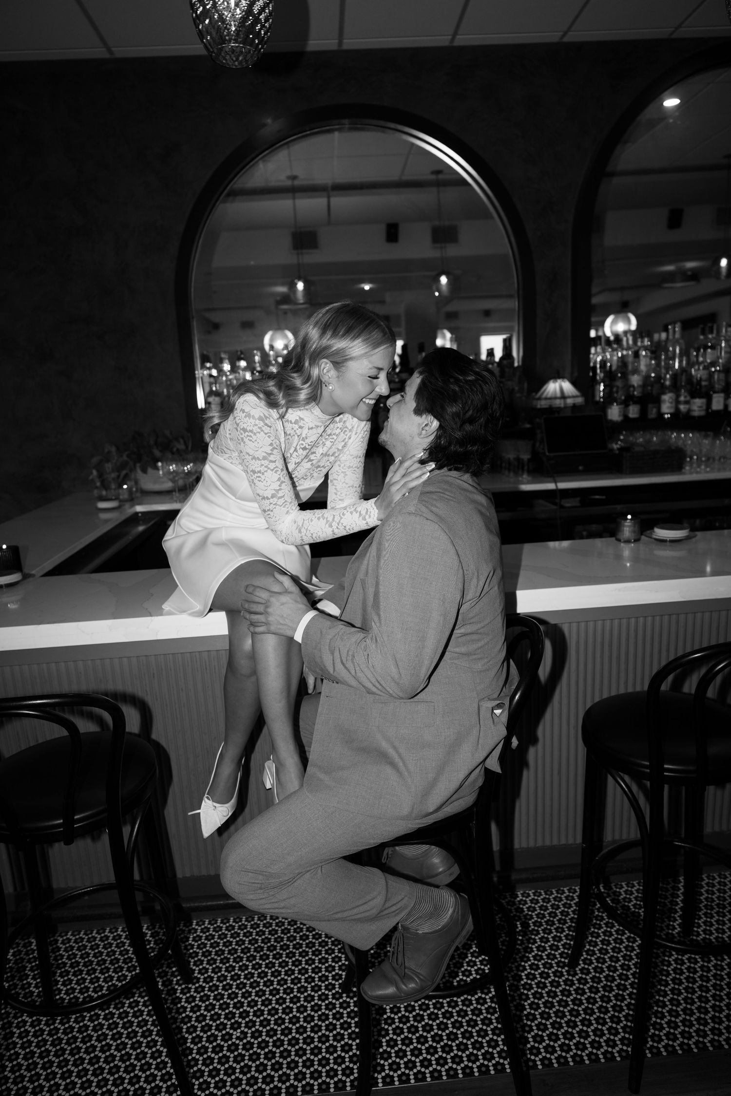 Black and white photo of a bride in a lace dress sitting on a bar counter, nose to nose with her groom on a stool.