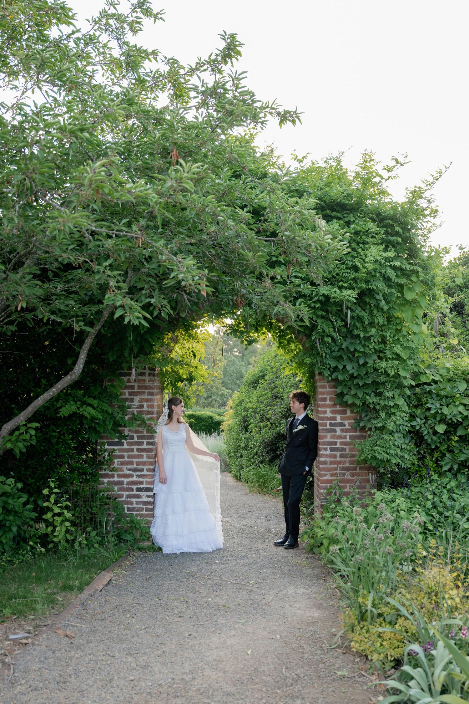 Bride and groom stand facing each other under a lush green garden archway at their wedding.
