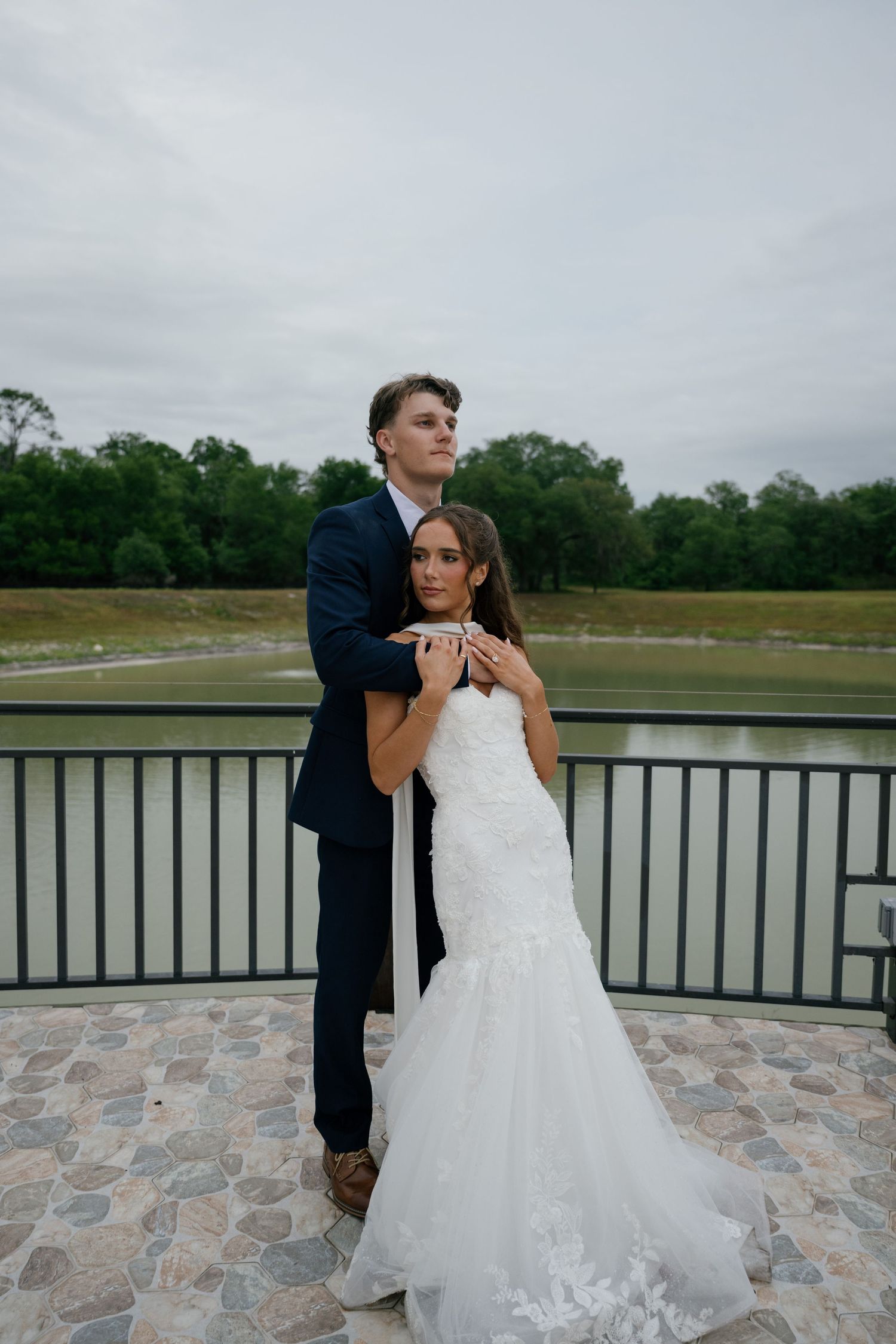 Bride and groom embrace on a stone terrace overlooking a pond, wearing a white lace gown and navy suit.