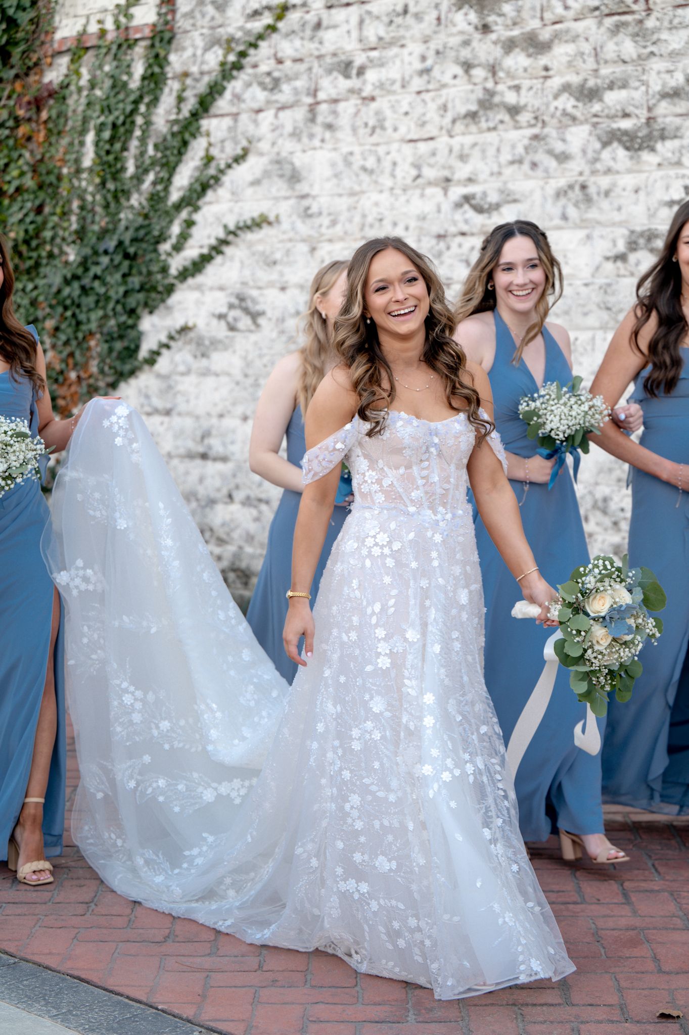 Smiling bride in floral lace wedding gown walks with bridesmaids in dusty blue dresses holding bouquets.