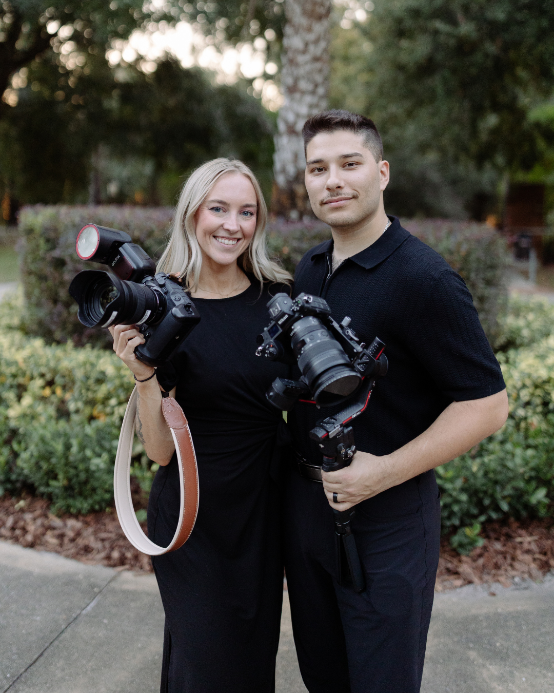 Couple in matching black outfits pose with professional cameras during an outdoor photography session.