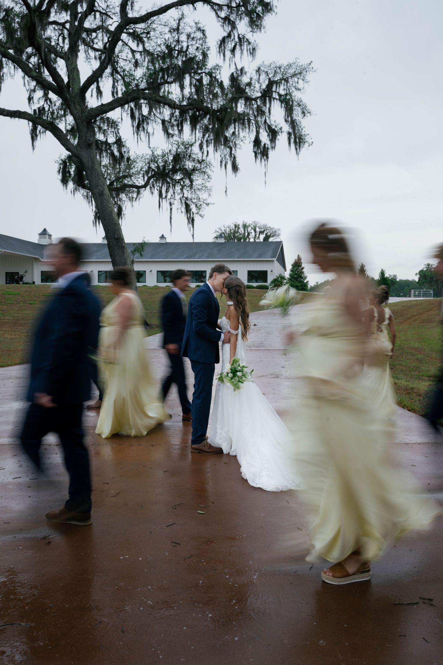 Bride and groom share a kiss surrounded by blurred wedding party in yellow dresses and navy suits on a rainy day.