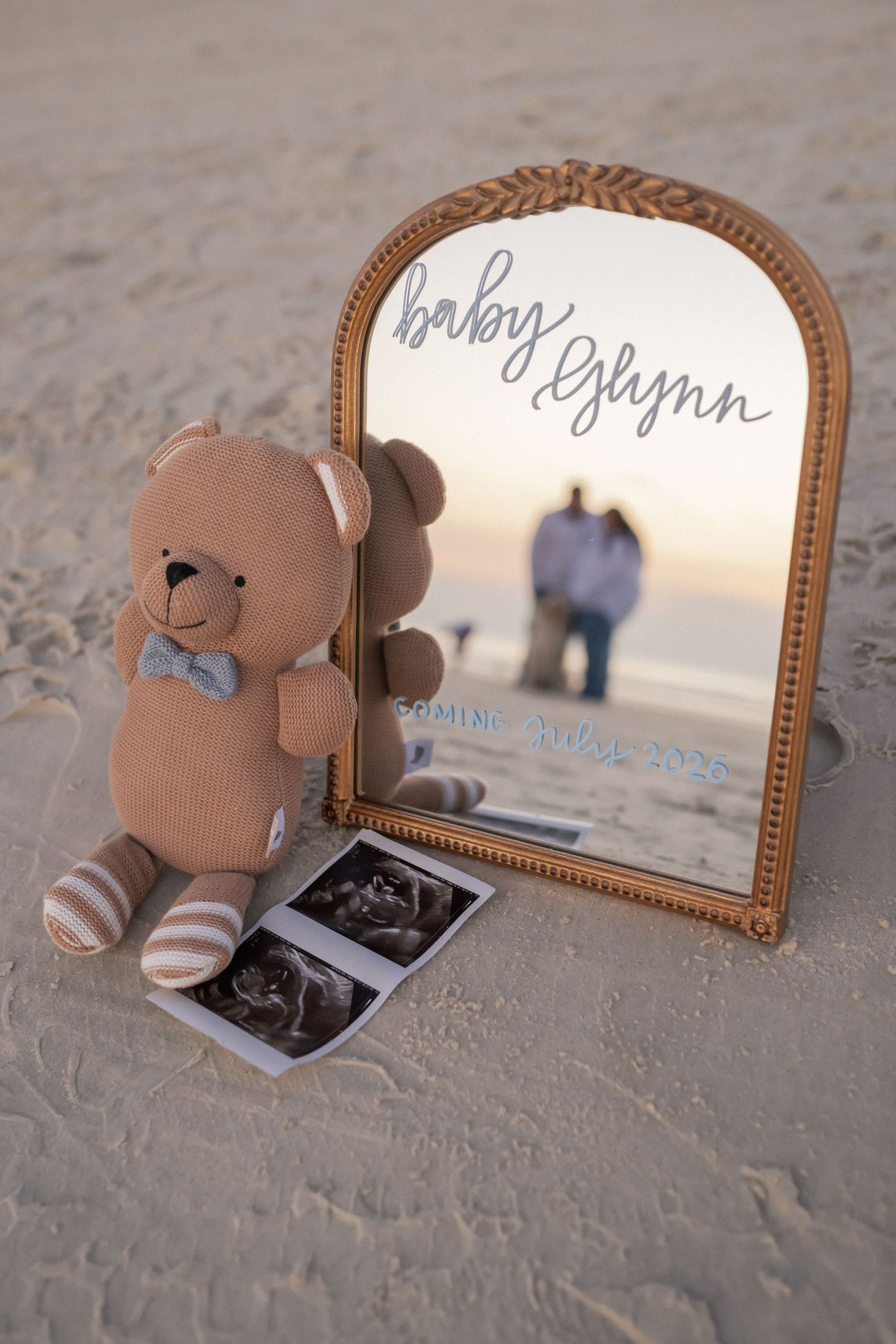 Teddy bear with bow tie beside gold mirror on beach announcing Baby Glynn coming 2025 with ultrasound photos.