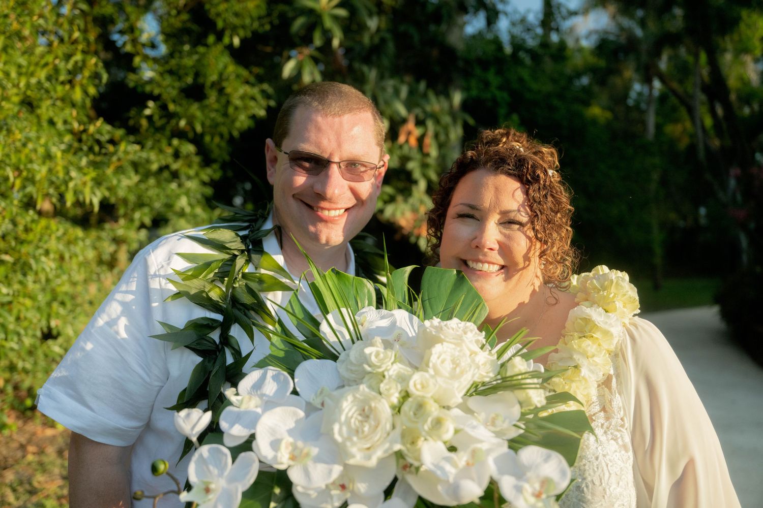 Happy couple smiling together outdoors, bride holding white floral bouquet with orchids and roses, lush green trees in background.