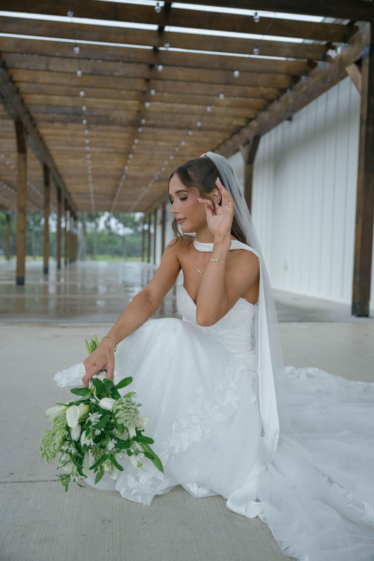 Bride in white gown and veil sits gracefully holding white floral bouquet under rustic wooden pergola.