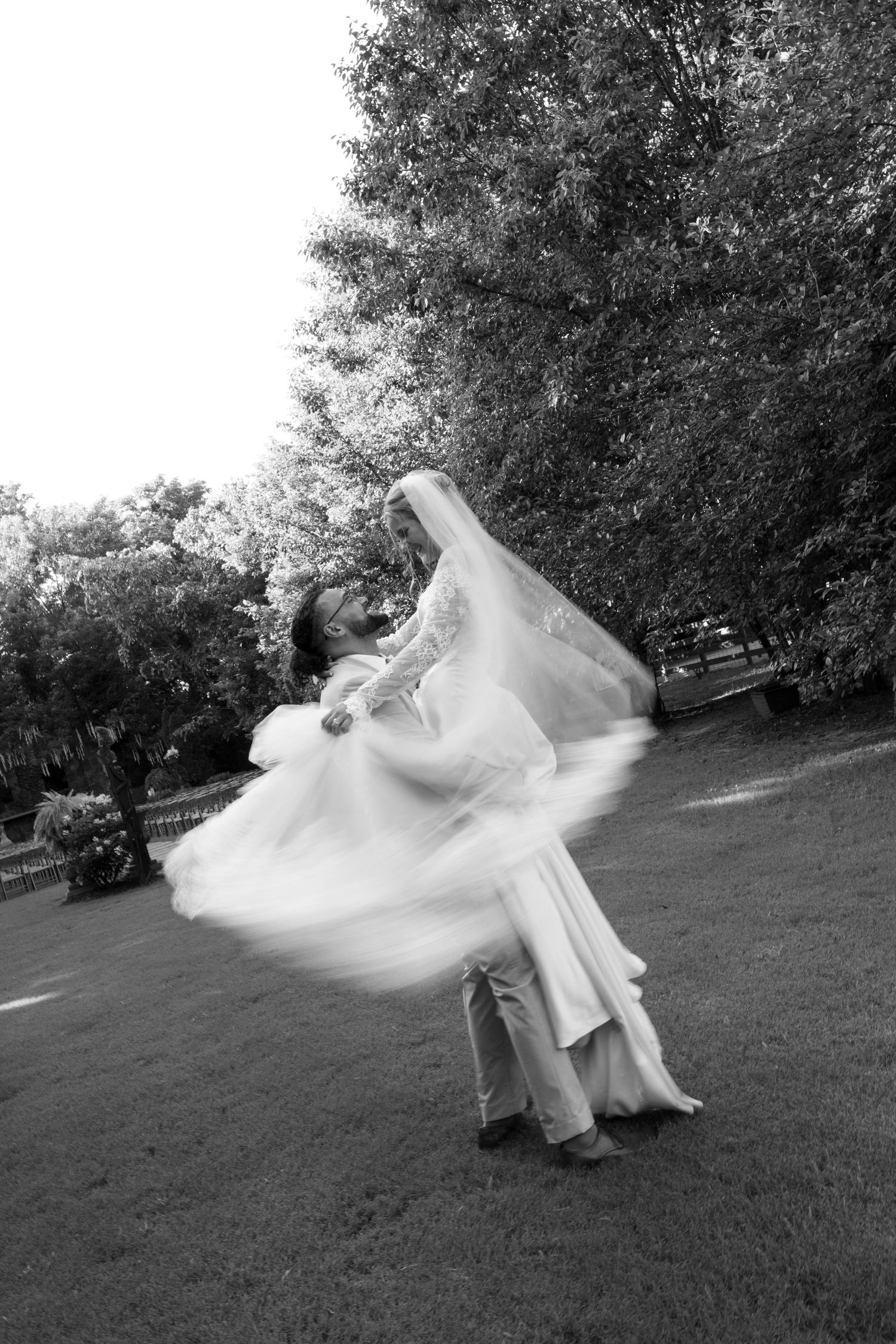 Black and white photo of groom spinning bride in wedding dress outdoors, veil flowing, trees in background.