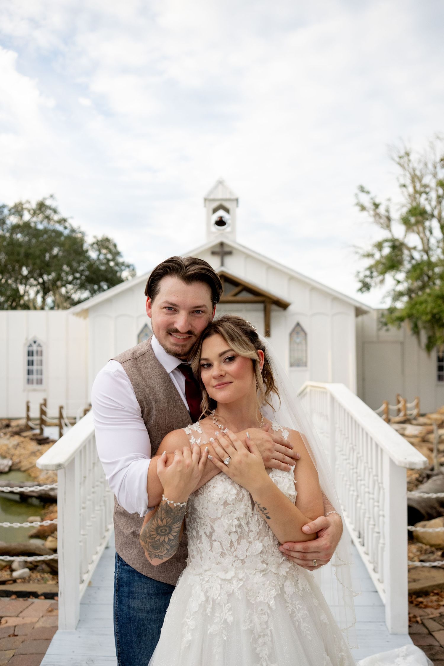 Newlyweds pose lovingly in front of a white church while showing off wedding rings.