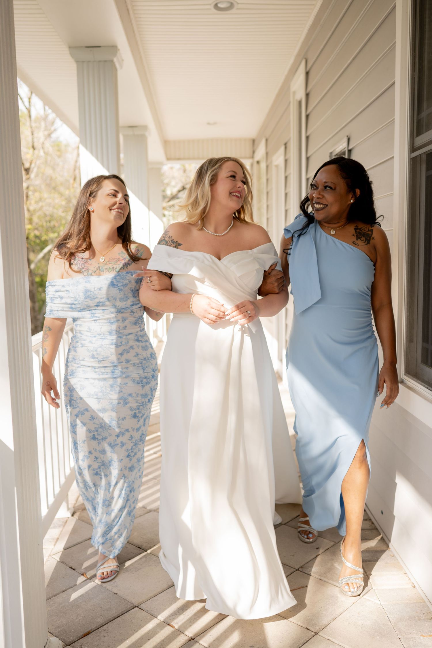 Bridesmaids in light blue and white dresses dance joyfully along a sunlit porch.