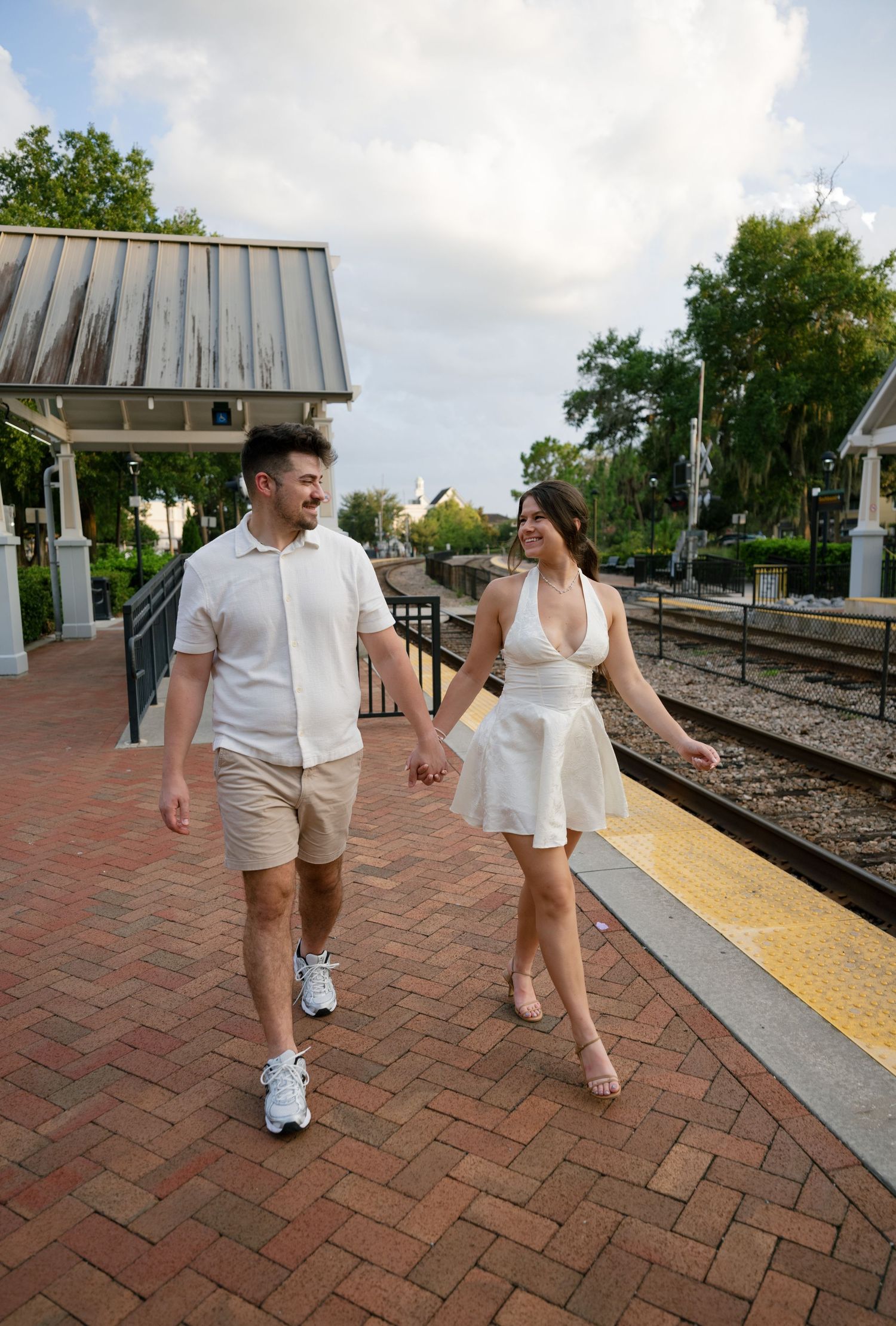 Couple holding hands and walking along a brick train station platform, dressed in white outfits on a cloudy day.