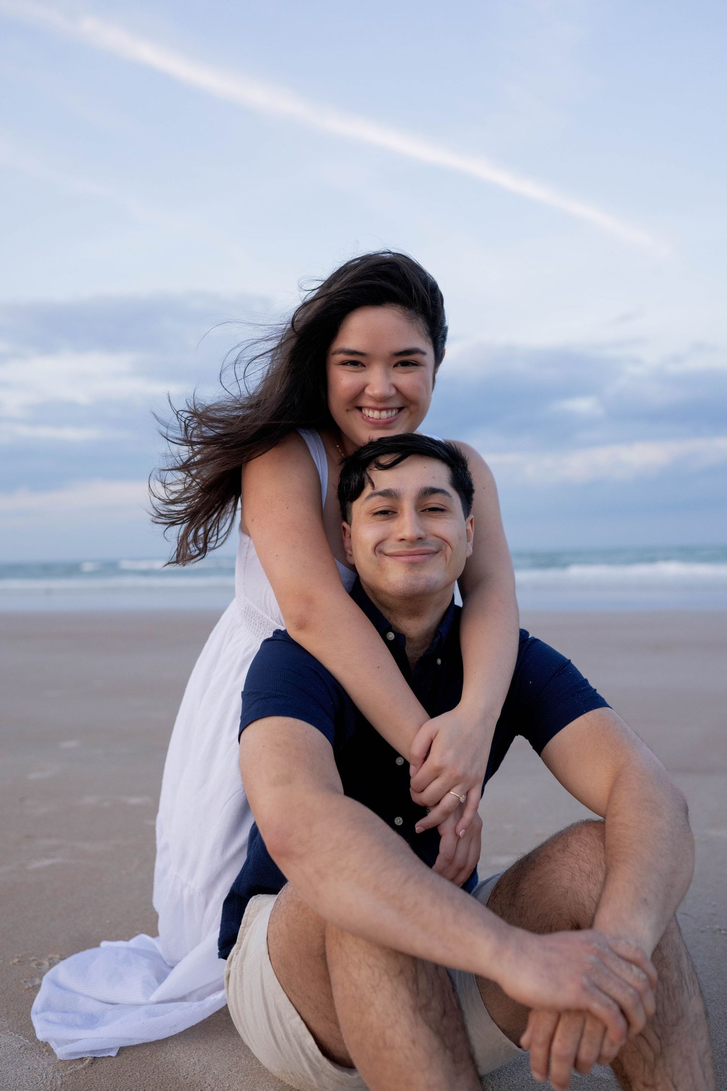 Couple embracing on a sandy beach at dusk, woman in white dress hugging man from behind as he sits smiling.