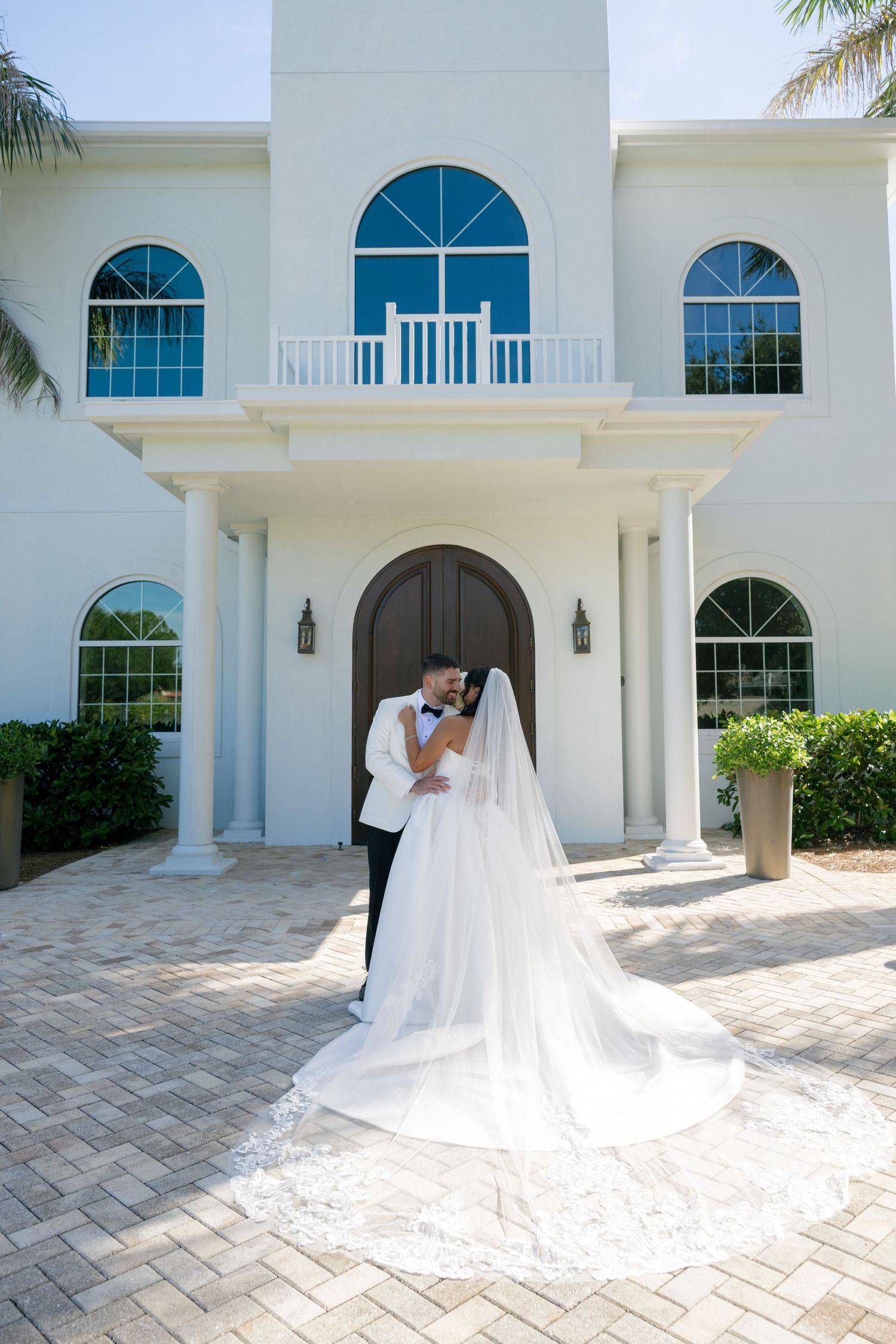 Bride and groom embrace on a brick paver driveway in front of a white mansion, her long lace-trimmed veil flowing behind her.