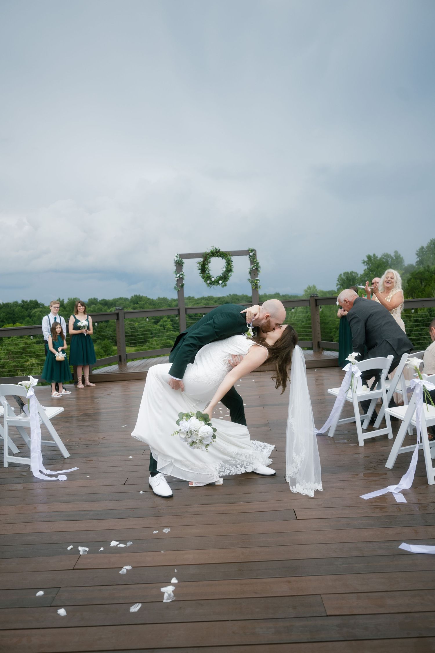 Groom dips bride for a kiss at outdoor wedding ceremony on wooden deck with green wreath arch and white chairs.