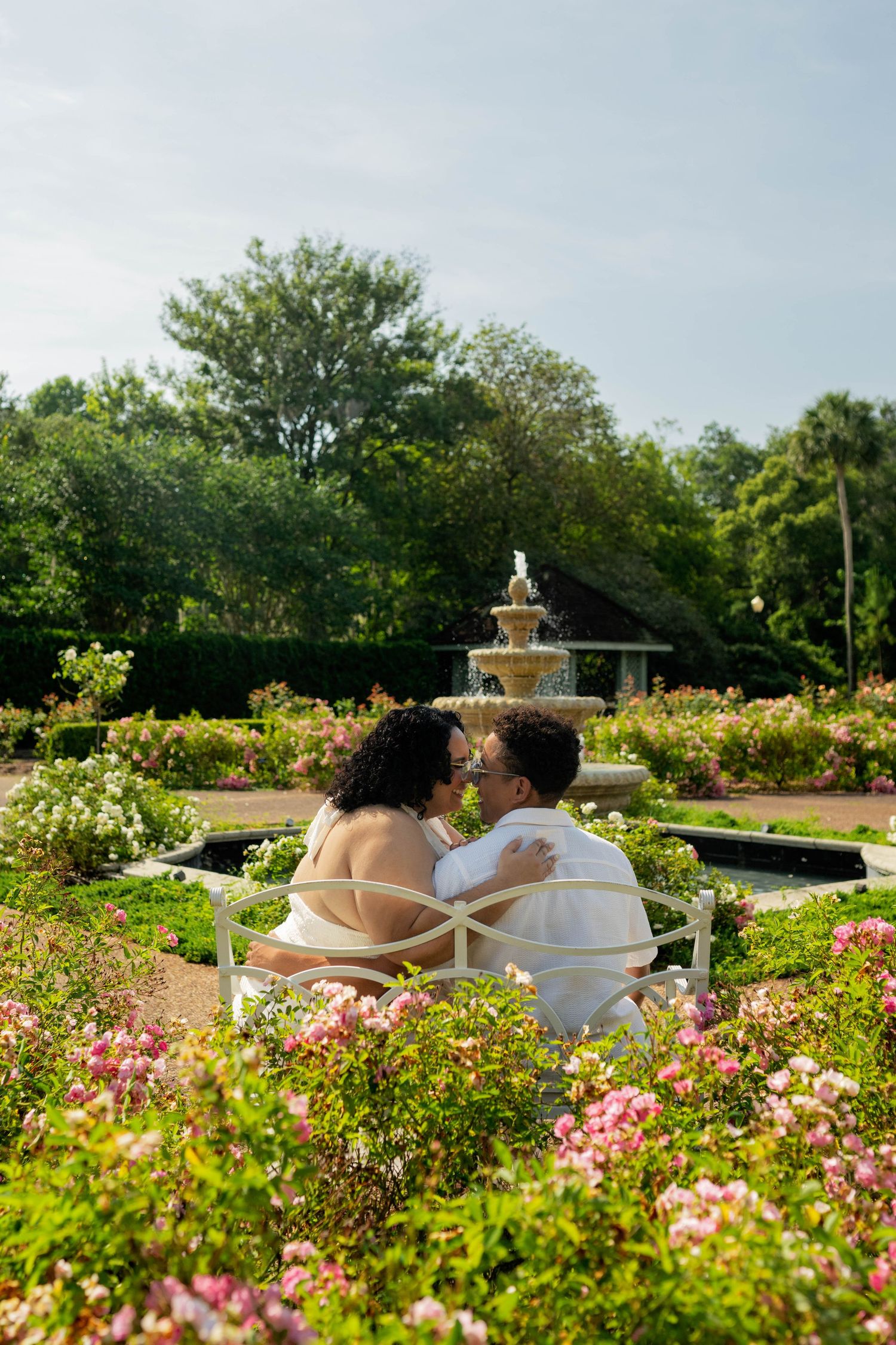Couple sitting on a bench in a romantic garden with a fountain, surrounded by colorful flowers and lush greenery.