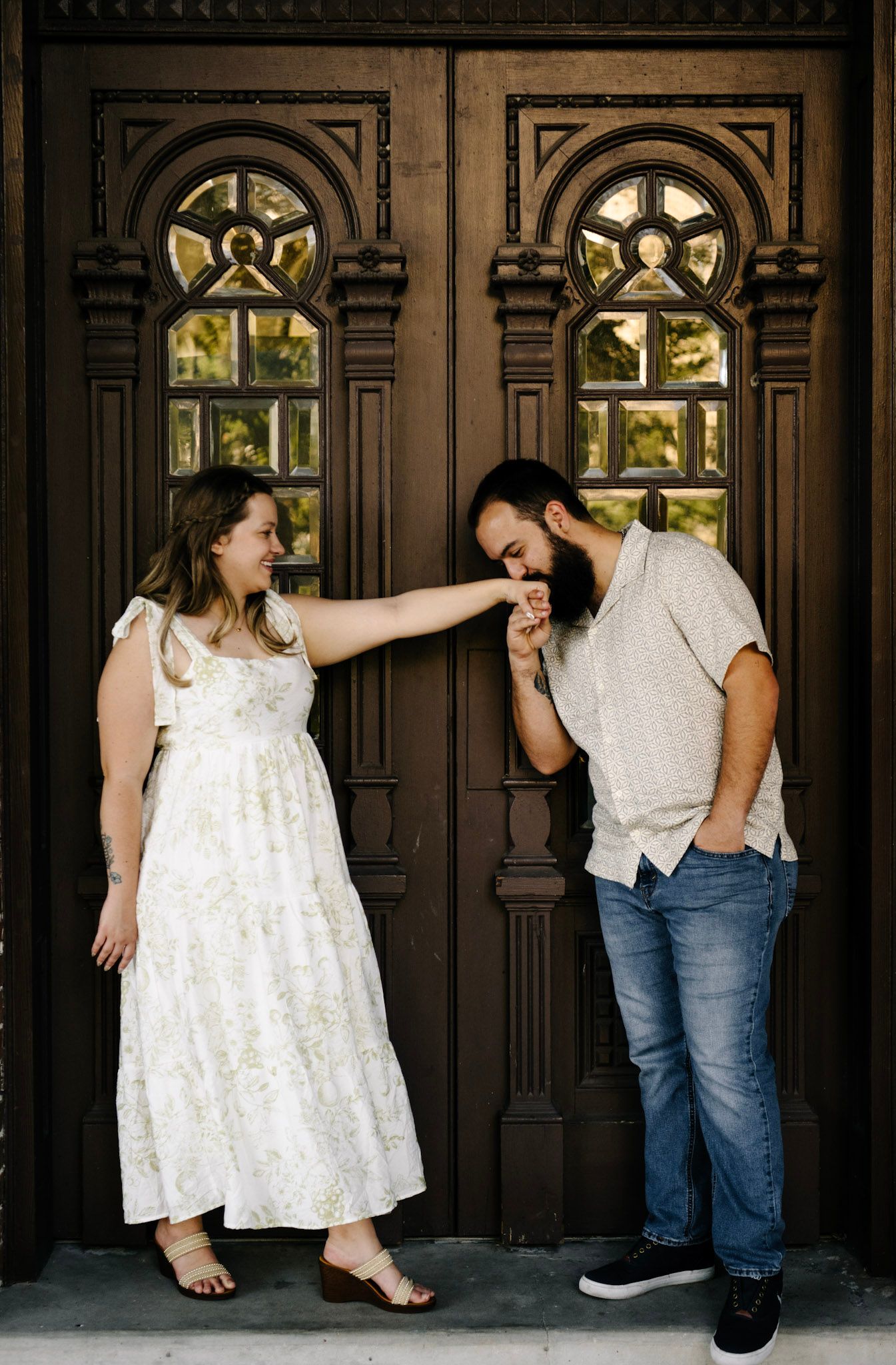 A romantic couple poses playfully in front of ornate wooden doors with arched windows.