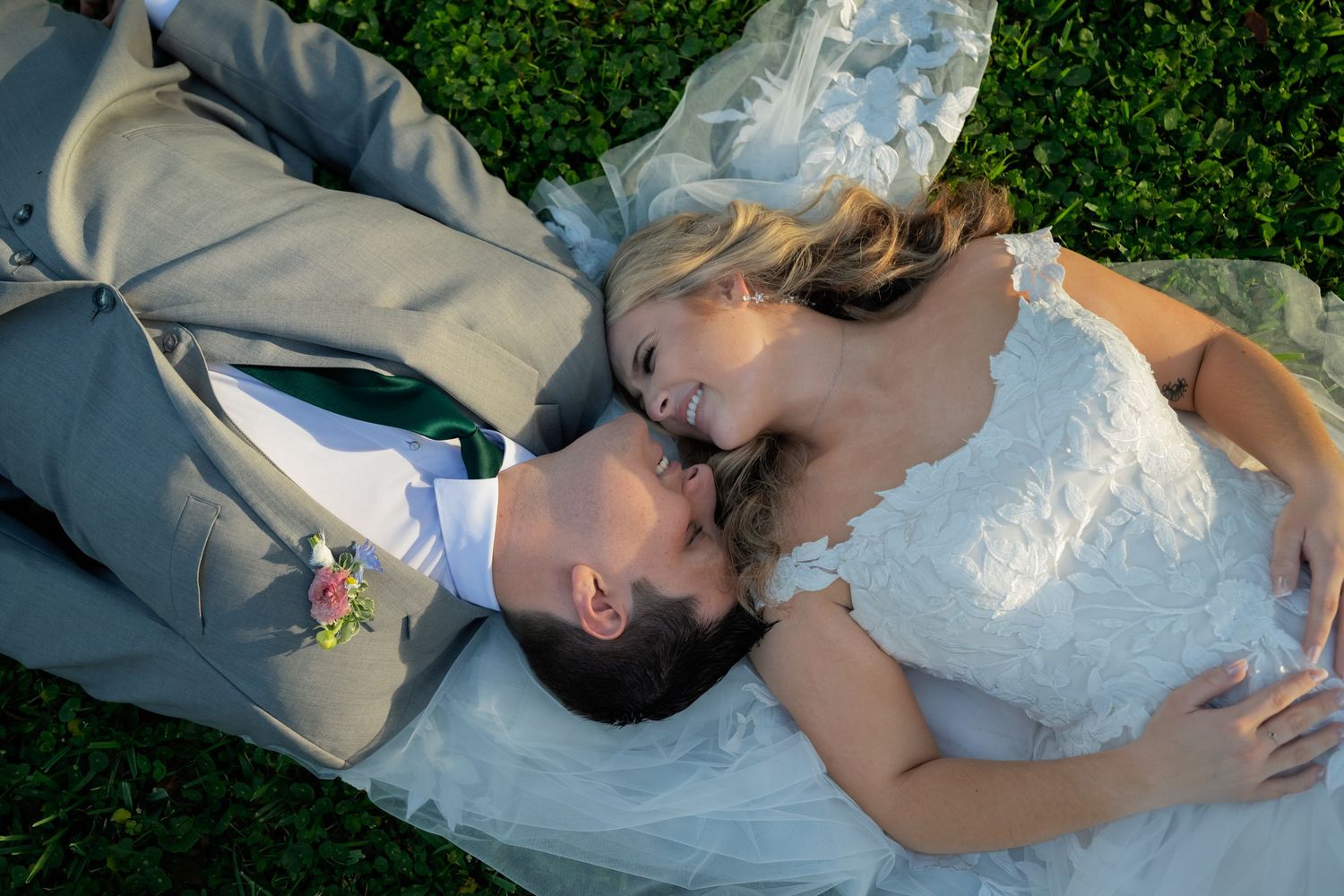 A couple in wedding attire lying on green grass in a romantic pose.