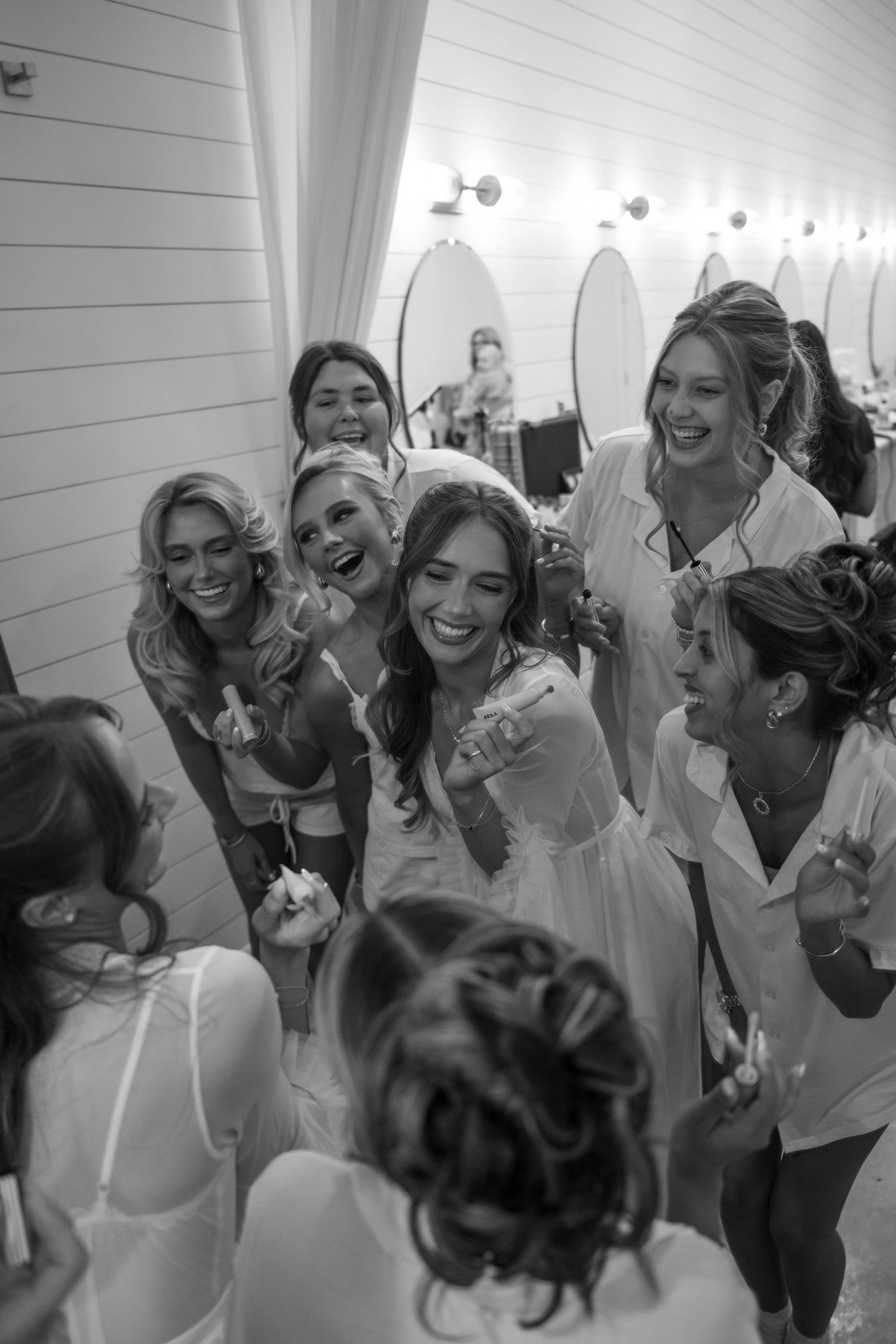 Bride laughs with bridesmaids in robes during wedding preparations in black and white photo.