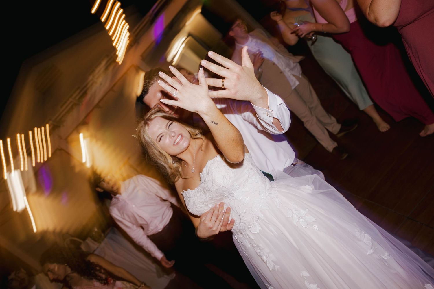 Bride in white wedding dress dancing joyfully at reception, hands raised toward camera with motion blur lights.