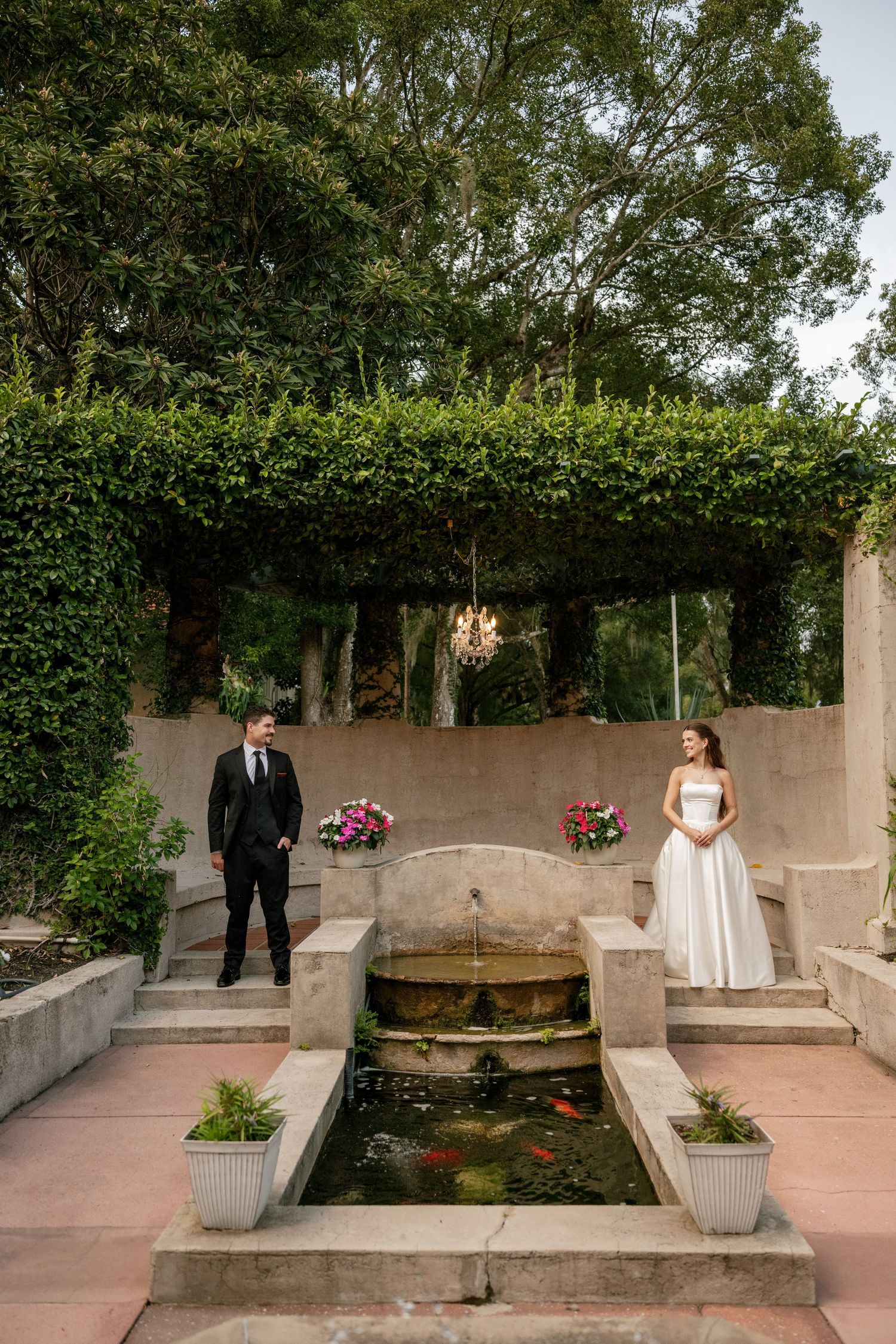 Bride in a white gown and groom in a black suit stand on opposite sides of a stone fountain and koi pond in a garden courtyard.