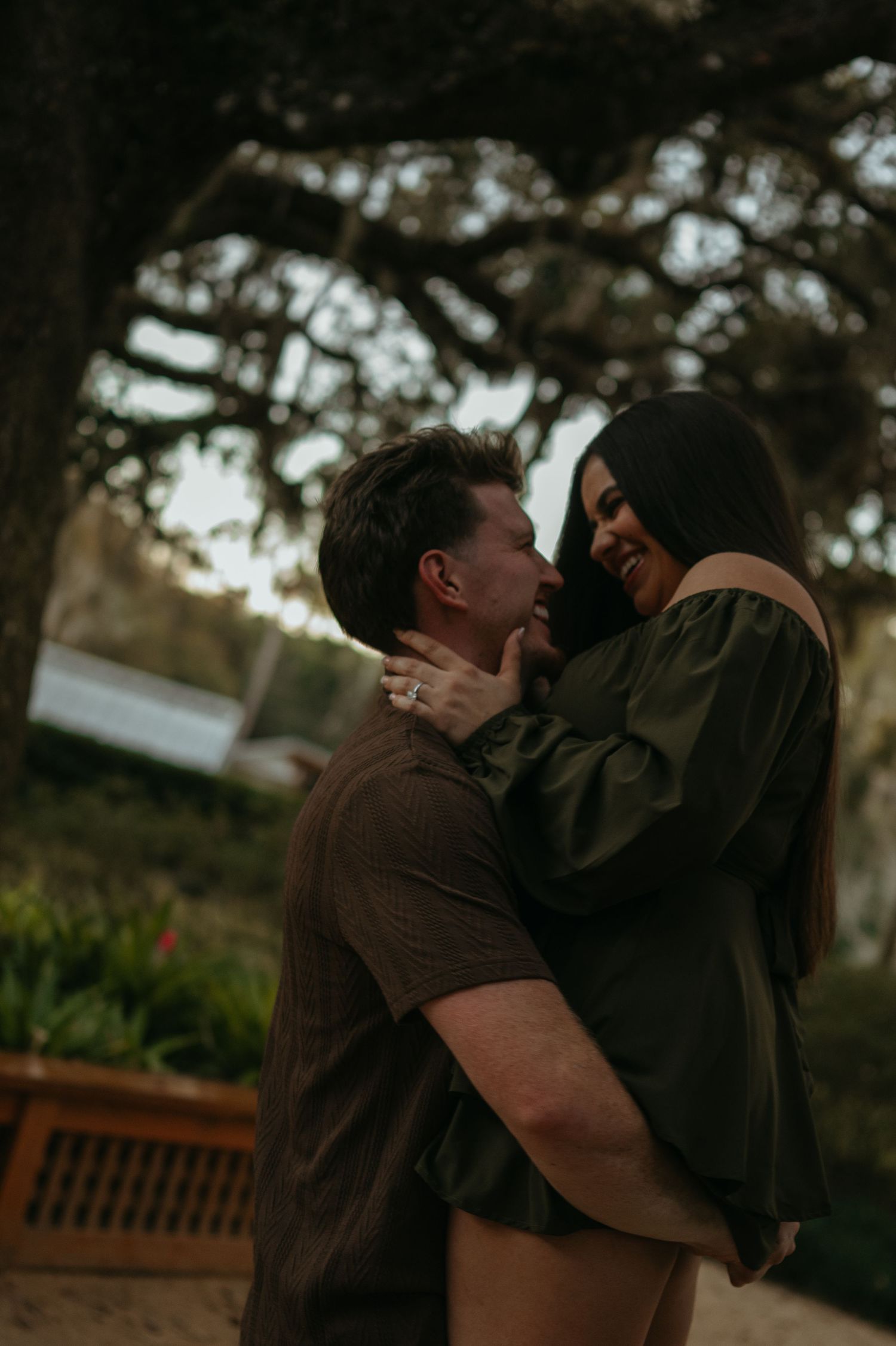 A romantic couple embraces in a garden at dusk, with trees and a wooden bench in the background.