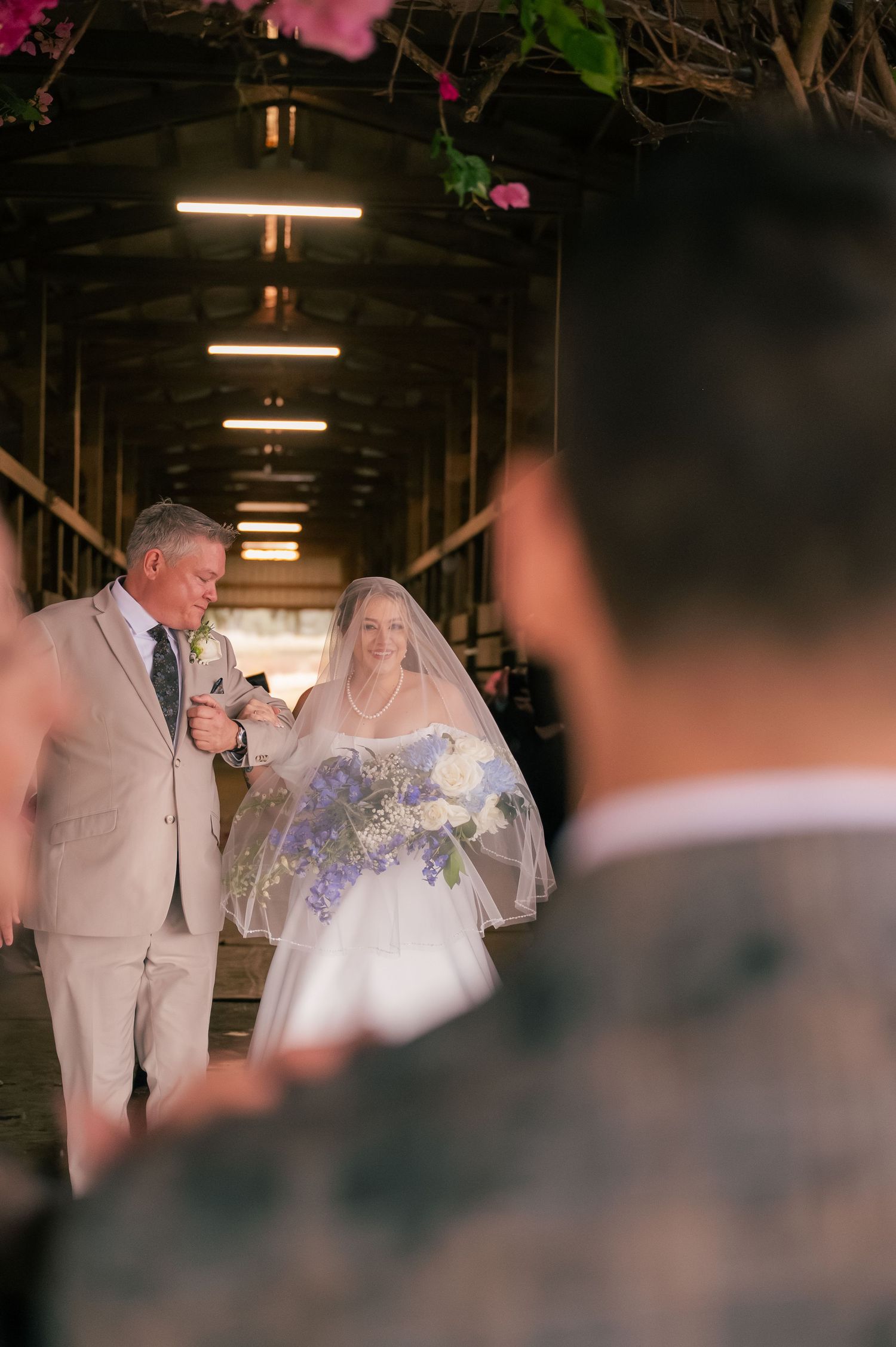 A couple walks down an illuminated covered walkway during a wedding ceremony with purple flowers in the bouquet.
