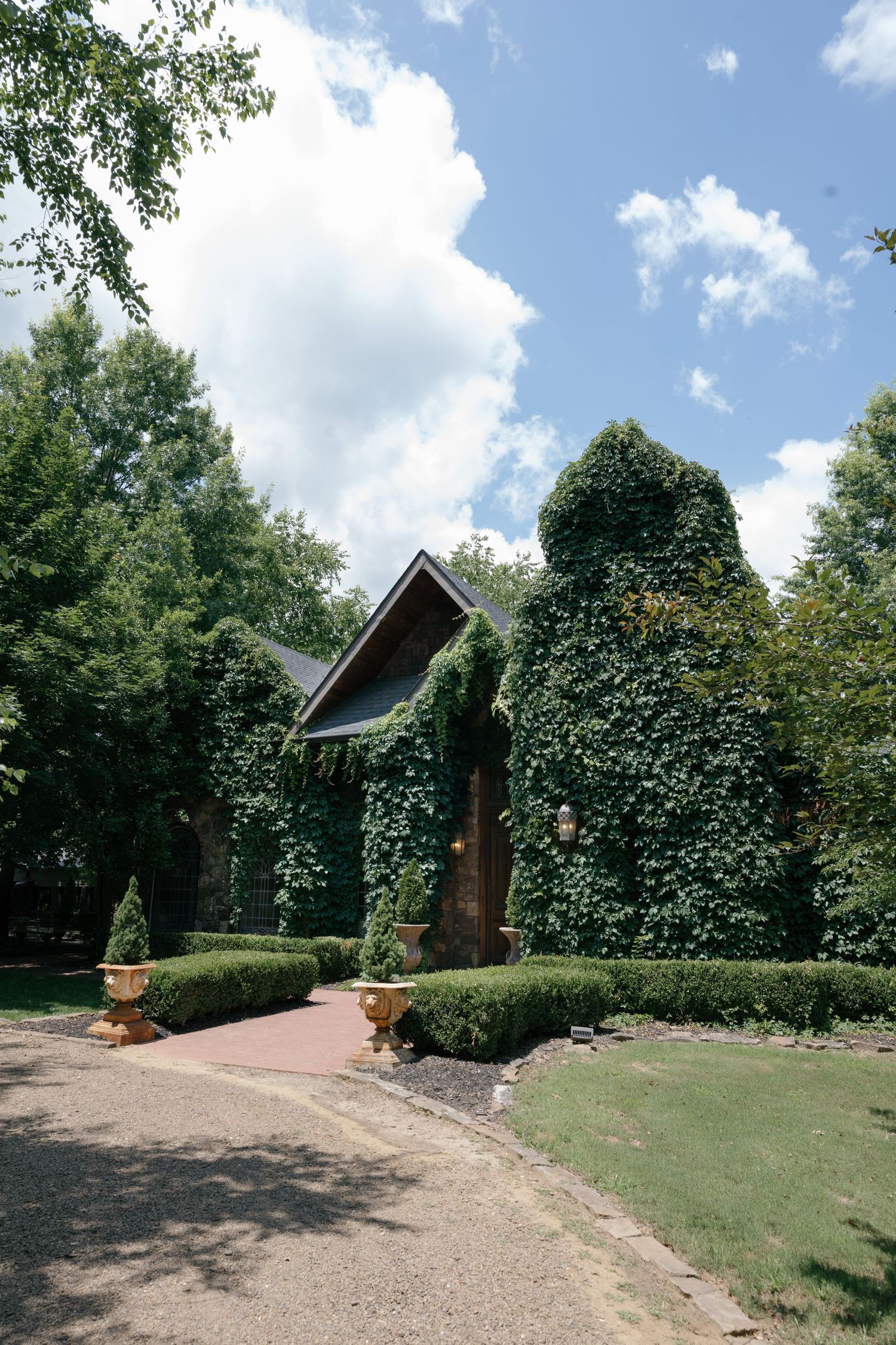 Ivy-covered chapel surrounded by lush green trees and manicured hedges with brick pathway and decorative urns.