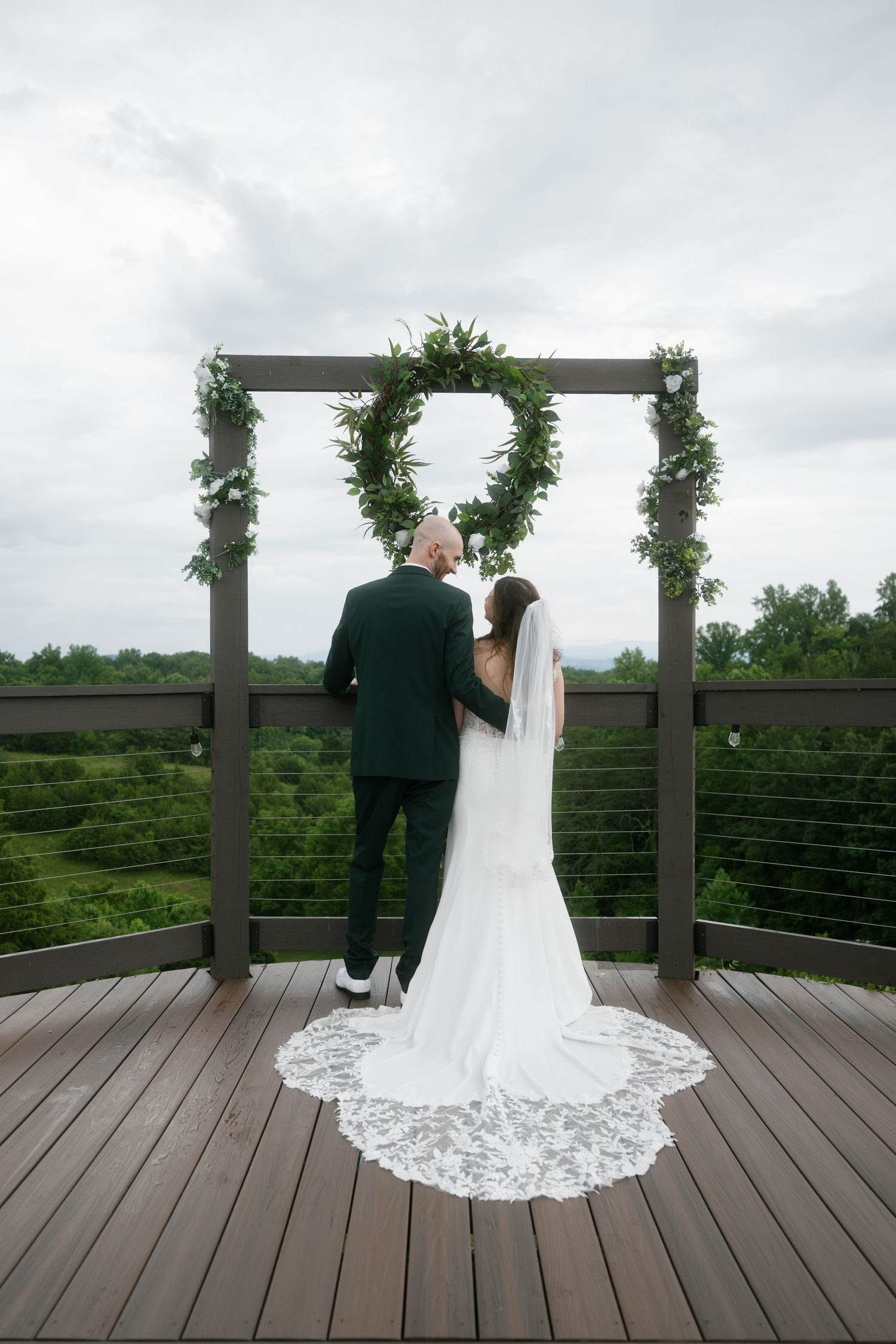 Bride and groom stand before a circular greenery arch on a wooden deck overlooking lush green hills on their wedding day.
