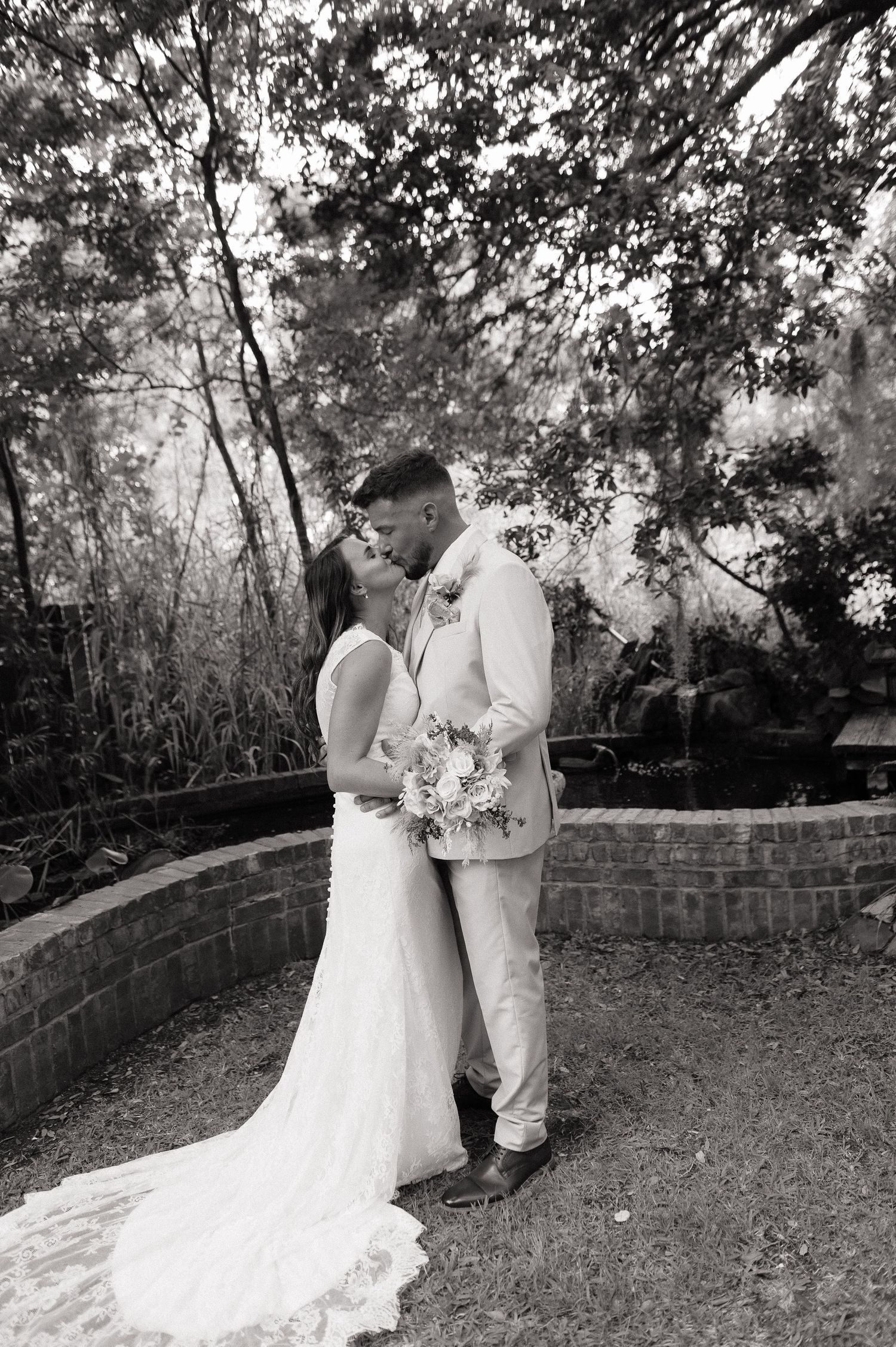Black and white photo of a bride and groom sharing a romantic kiss outdoors, surrounded by lush trees.