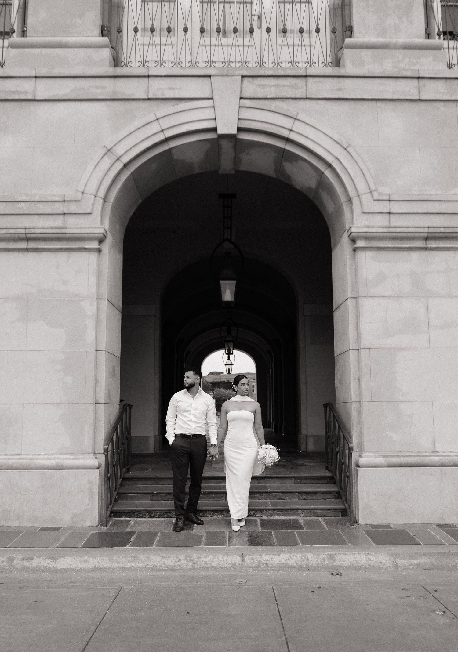 Black and white photo of a bride and groom walking through an elegant stone archway on their wedding day.