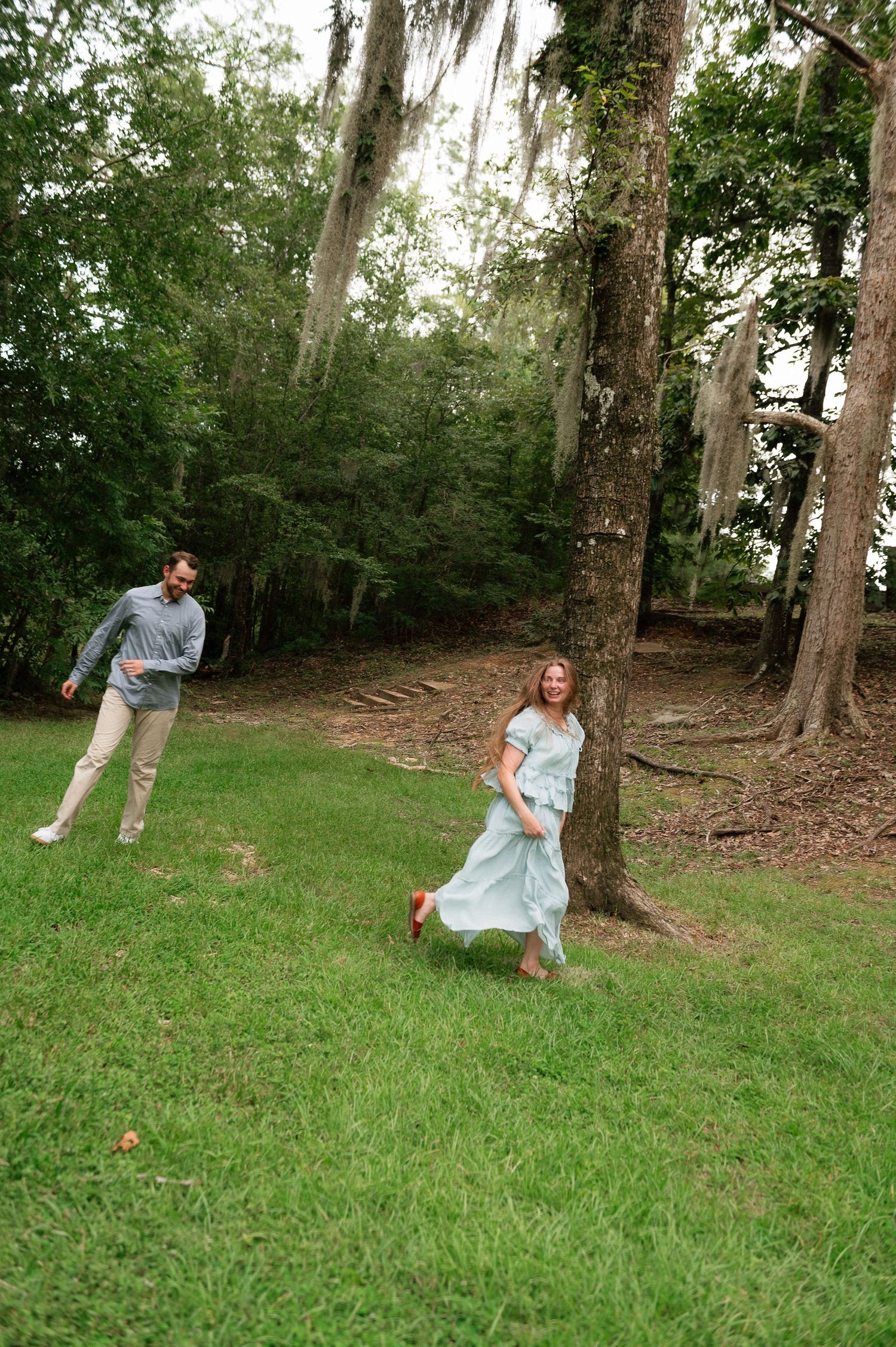A couple playfully chases each other around a large tree in a lush green park surrounded by moss-draped trees.