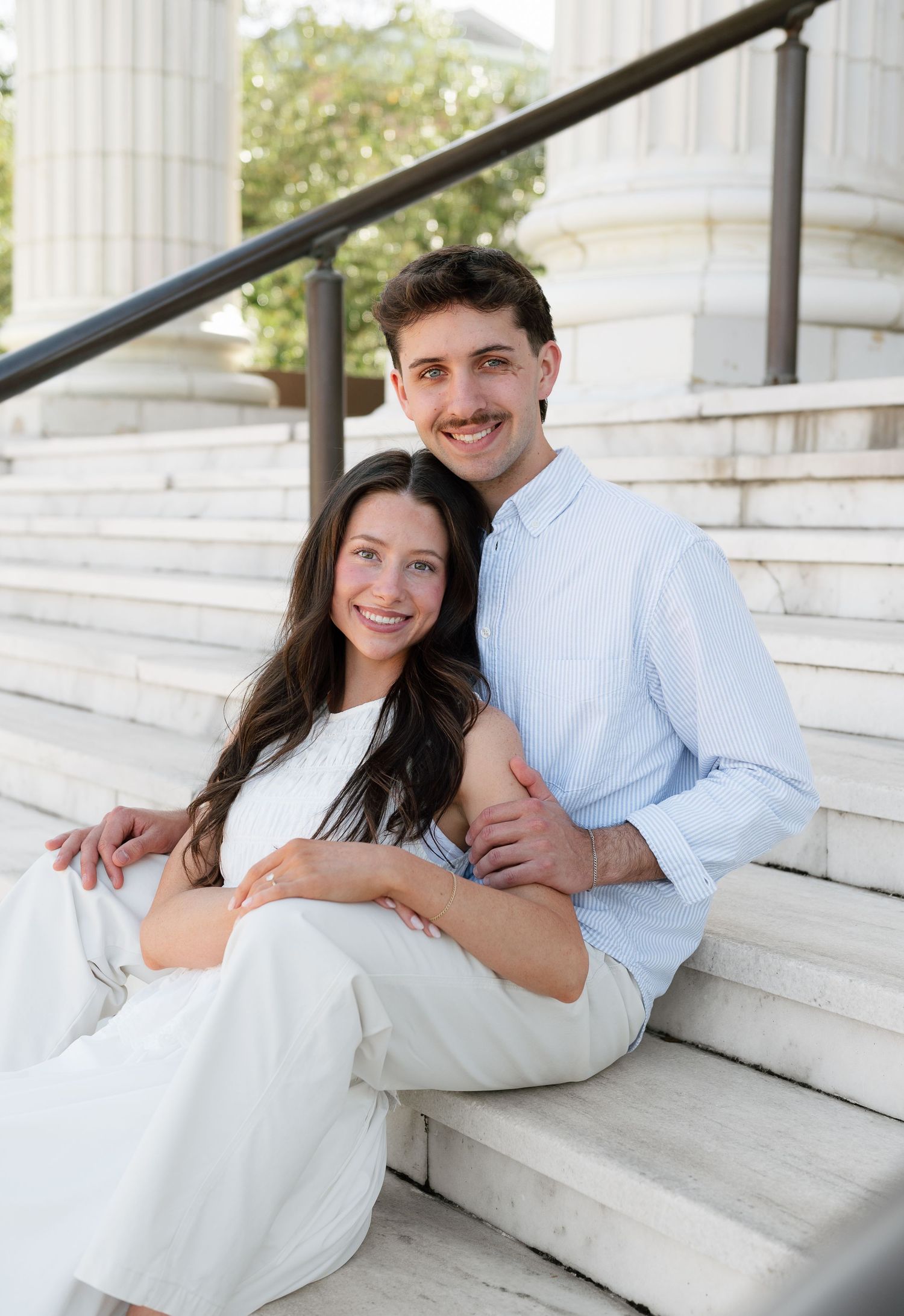 Smiling couple sitting on marble steps of a classical building, woman in white dress leaning against man in light blue shirt.